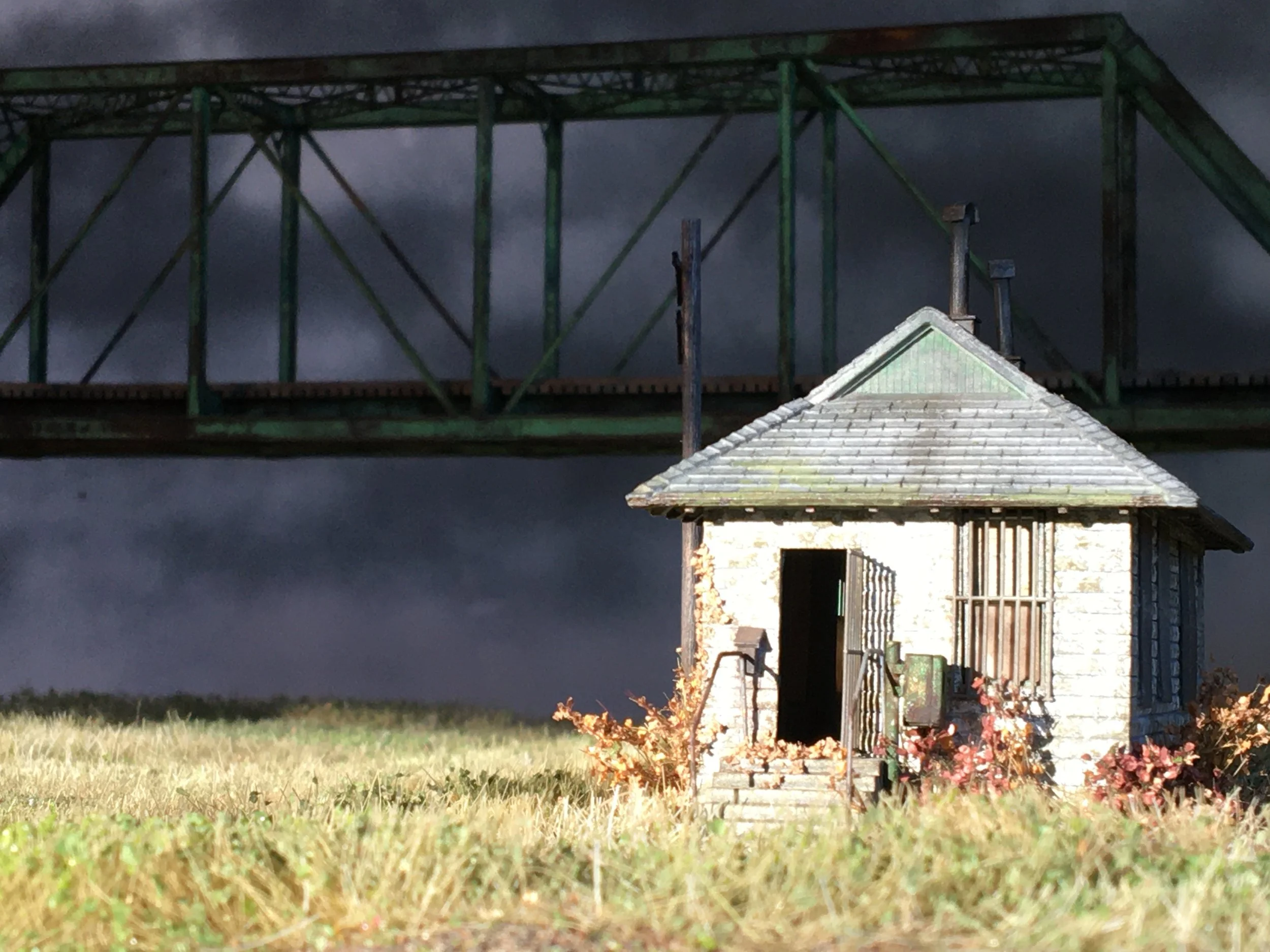 A small wooden house with a shingled roof, surrounded by grass and reddish shrubbery, with a large green metal bridge in the background under a dark, cloudy sky.