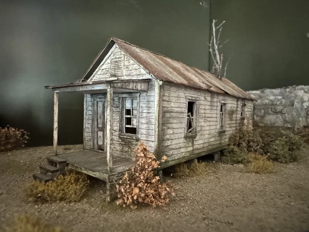 Model of an abandoned, weathered wooden house with a porch, missing window panes, surrounded by dry shrubs and a barren landscape.
