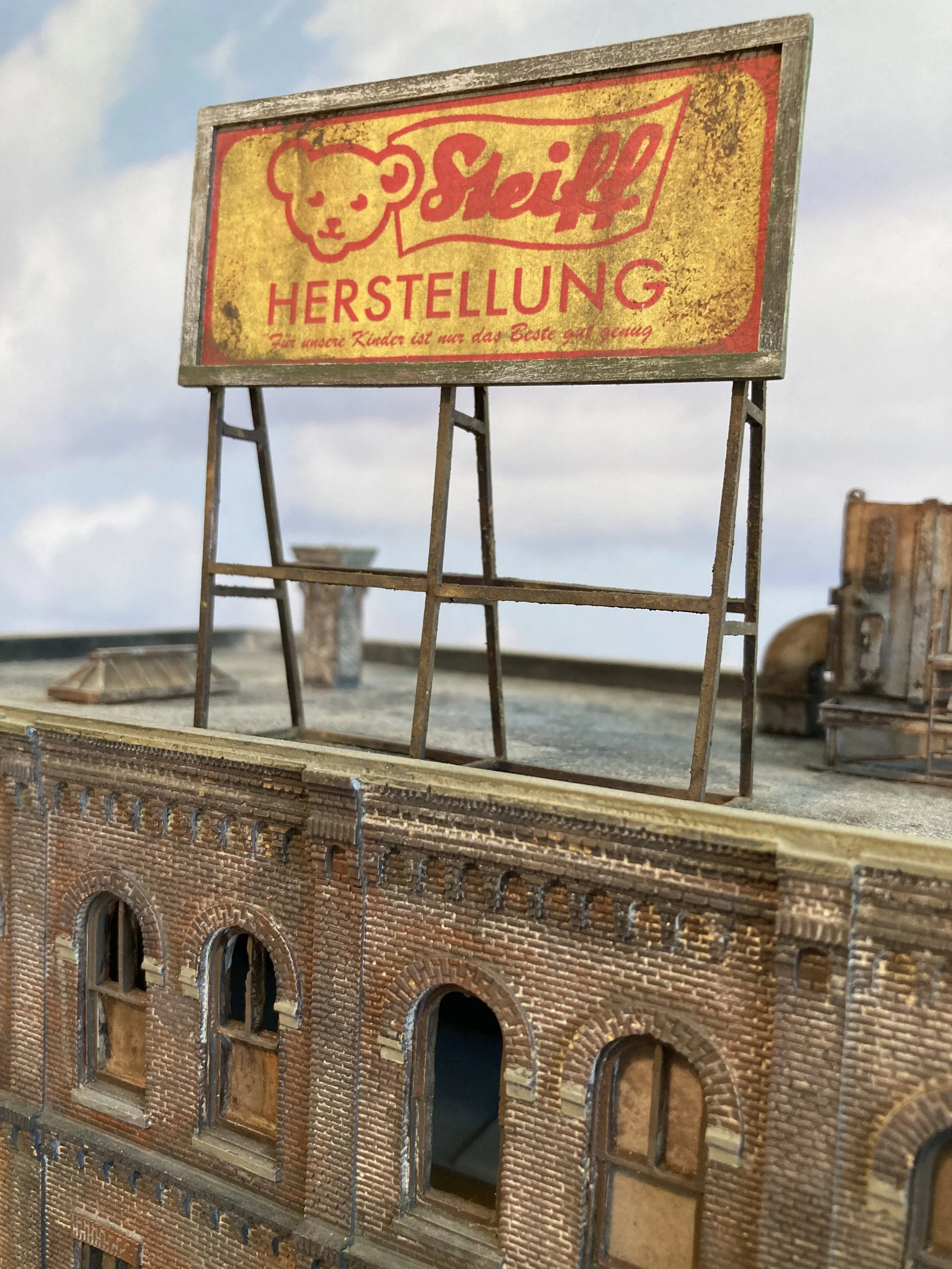 Close-up of a decorative building facade with detailed brickwork, and a red brick archway, possibly a miniature or model. In the background, there is a weathered sign with a bear logo, the word 'Shell' and German text about manufacturing.