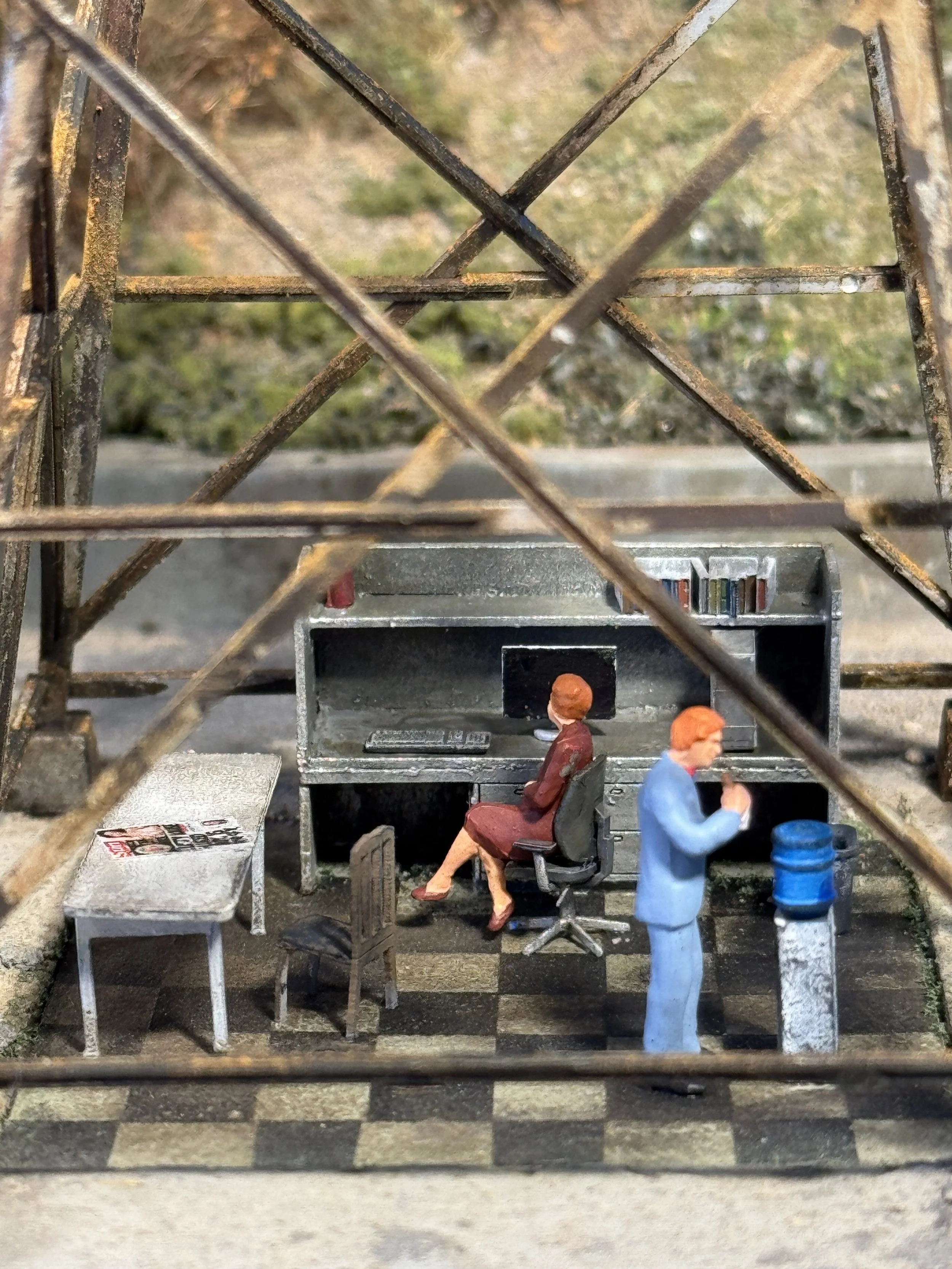 Miniature diorama of an office scene with two figurines of women, one sitting at a desk and the other standing, surrounded by office furniture and equipment, viewed through a metal framework.