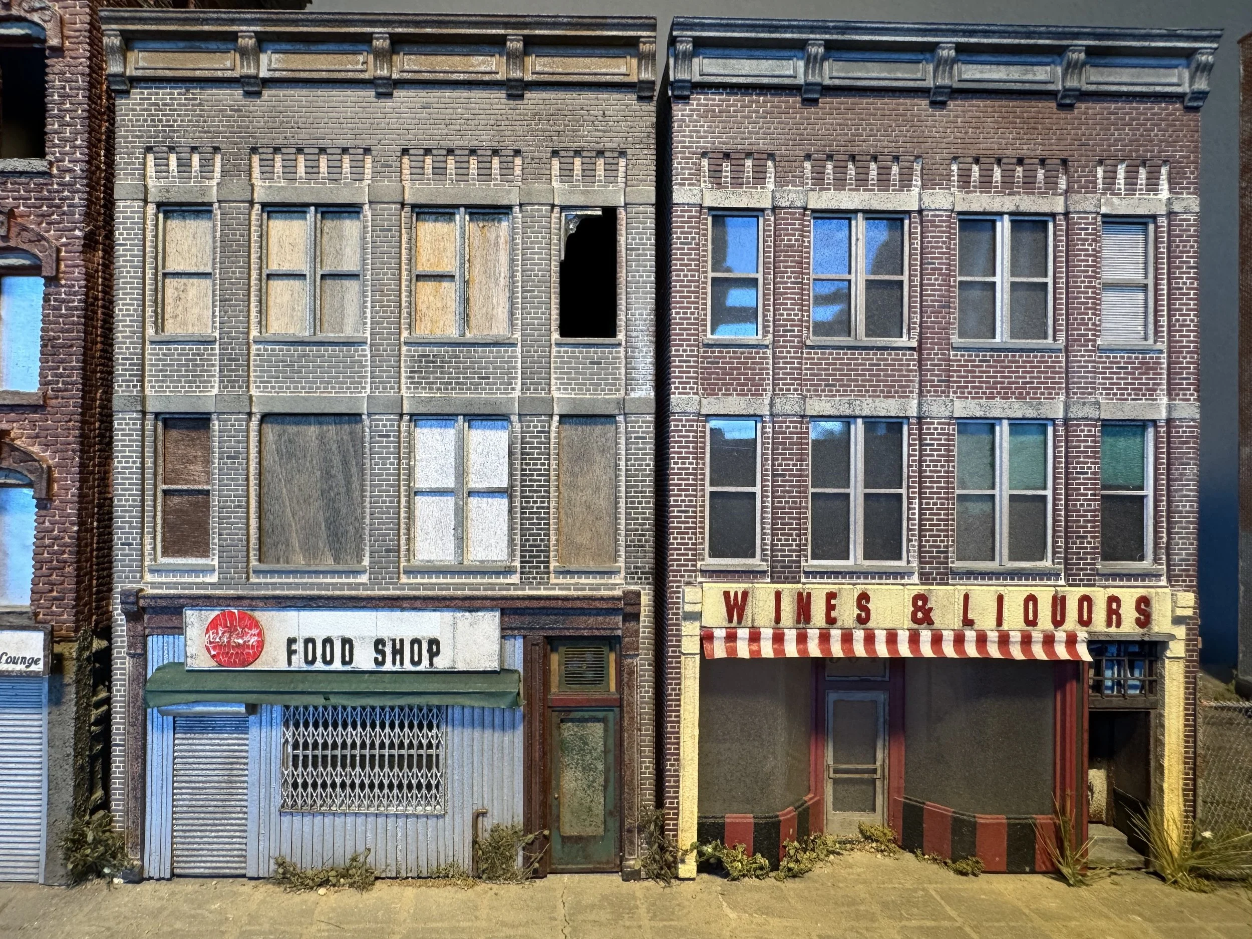 Three adjacent storefronts on a city street, two with boarded-up windows and one with a closed metal shutter. The left storefront has a green awning with a sign that reads 'FOOD SHOP,' and the right storefront has a yellow sign with red text that rea