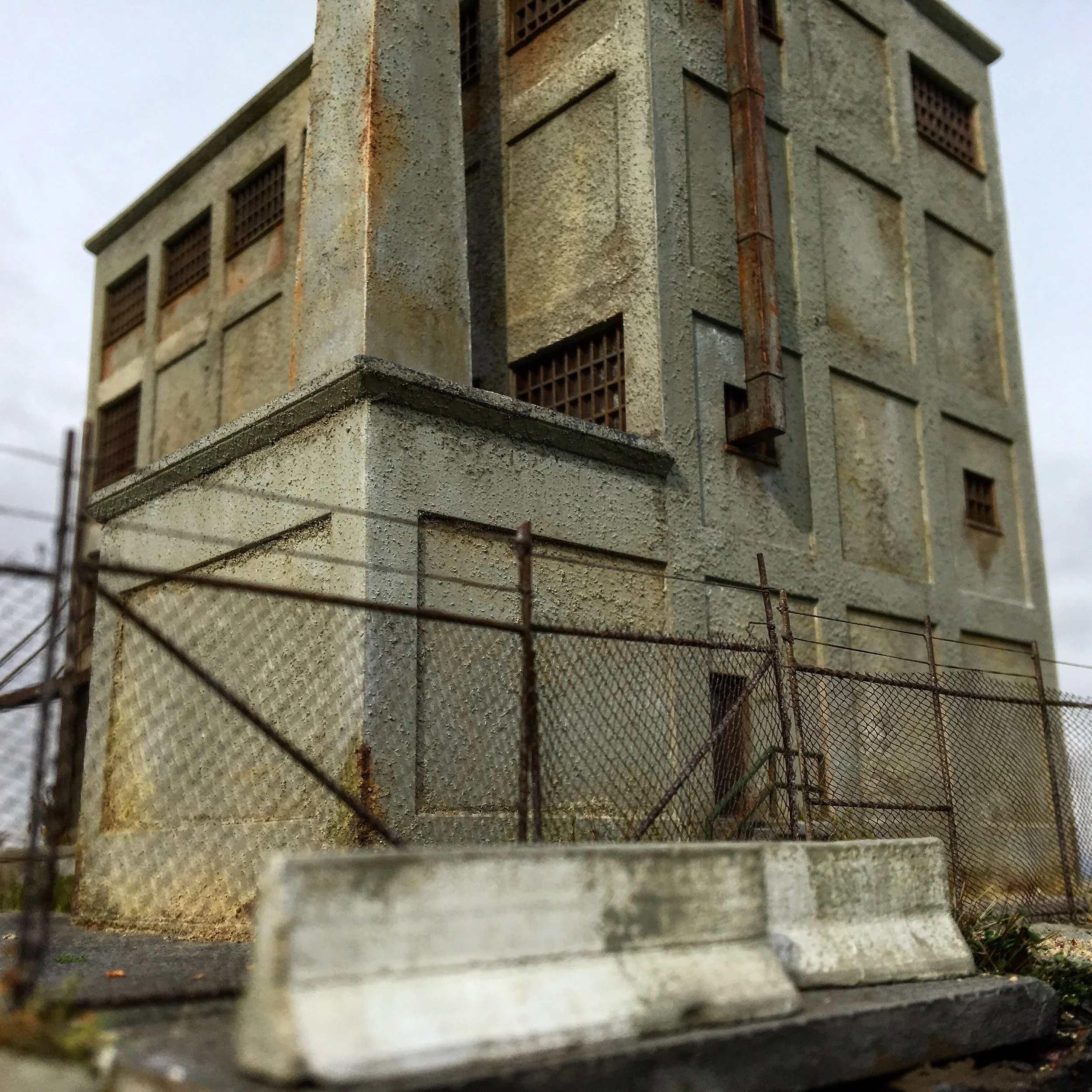 Close-up of a weathered building corner with a rusty drainpipe, seen through a rusted metal fence, cloudy sky in the background.