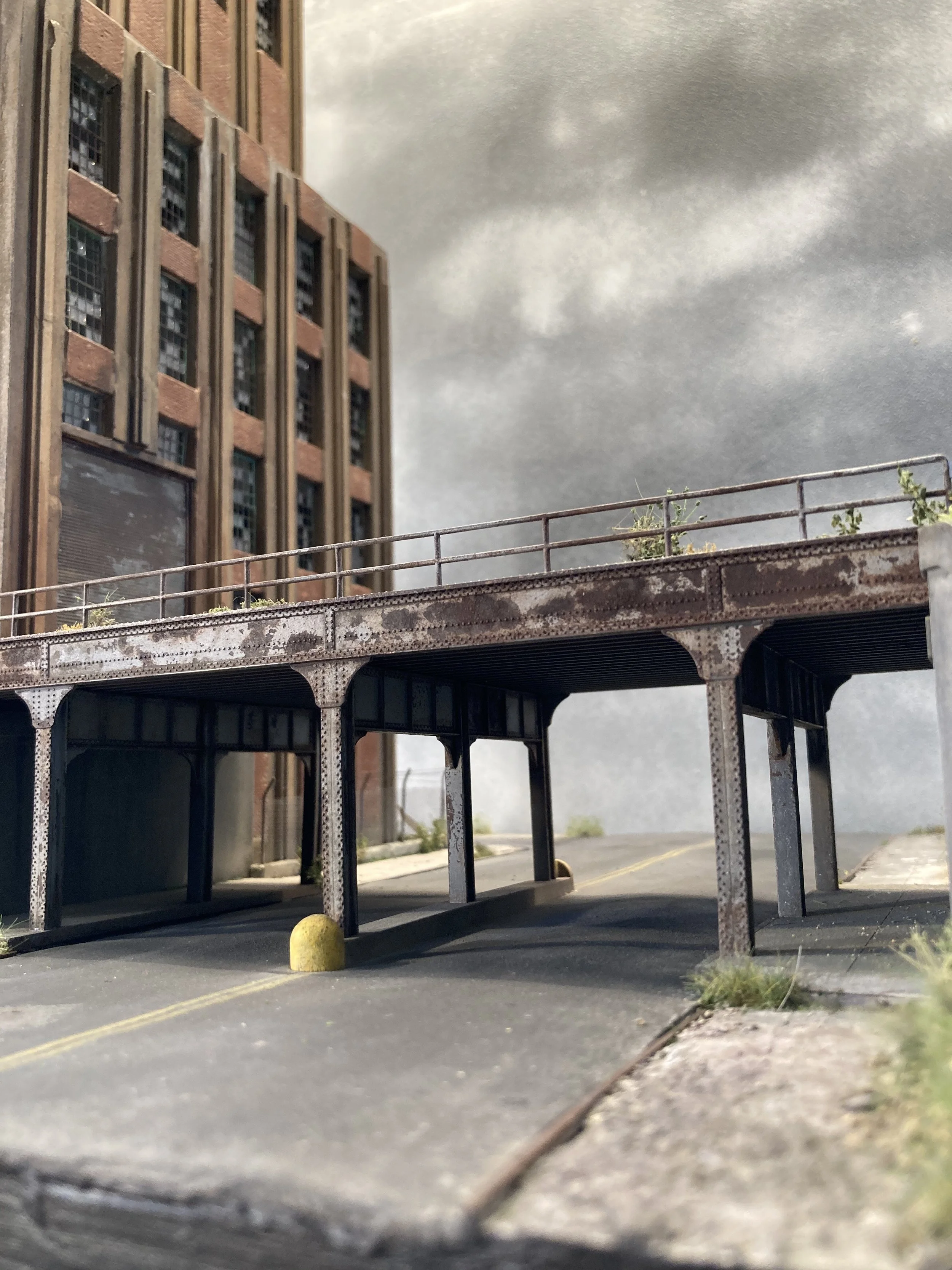 Miniature model of a city street scene with a tall brick building, a rusted metal bridge, and a yellow ball on the road.