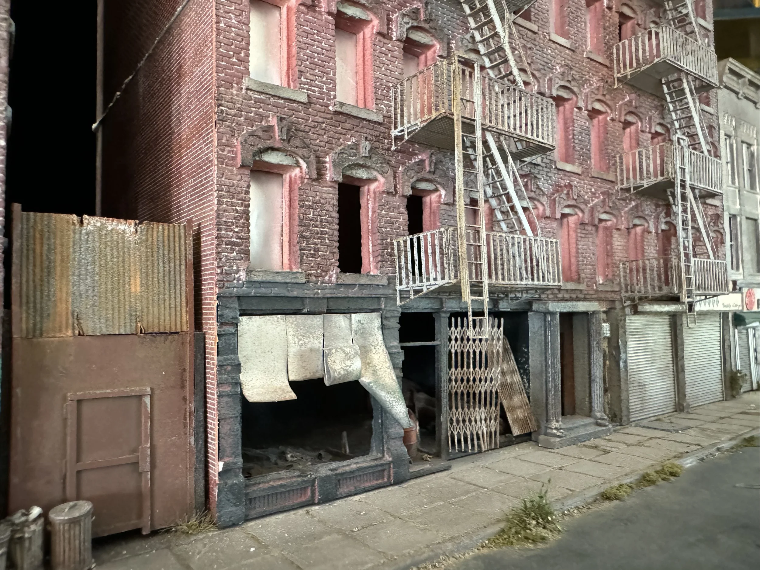 A detailed model of a multi-story brick building with fire escapes on the exterior, situated on a city sidewalk.