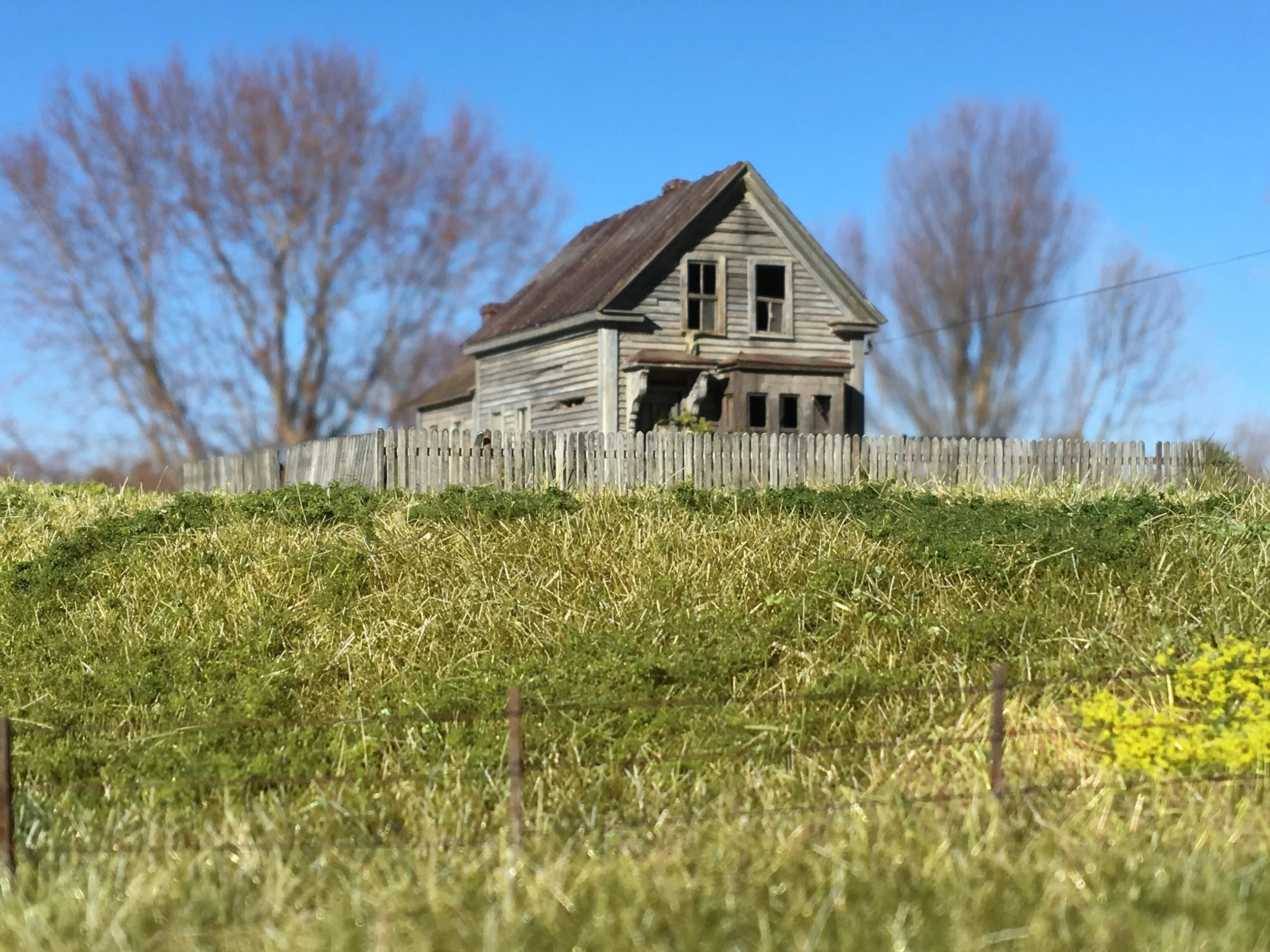 An old, weathered wooden house with a steeply pitched roof and broken windows, surrounded by leafless trees and a wooden fence, under a clear blue sky.