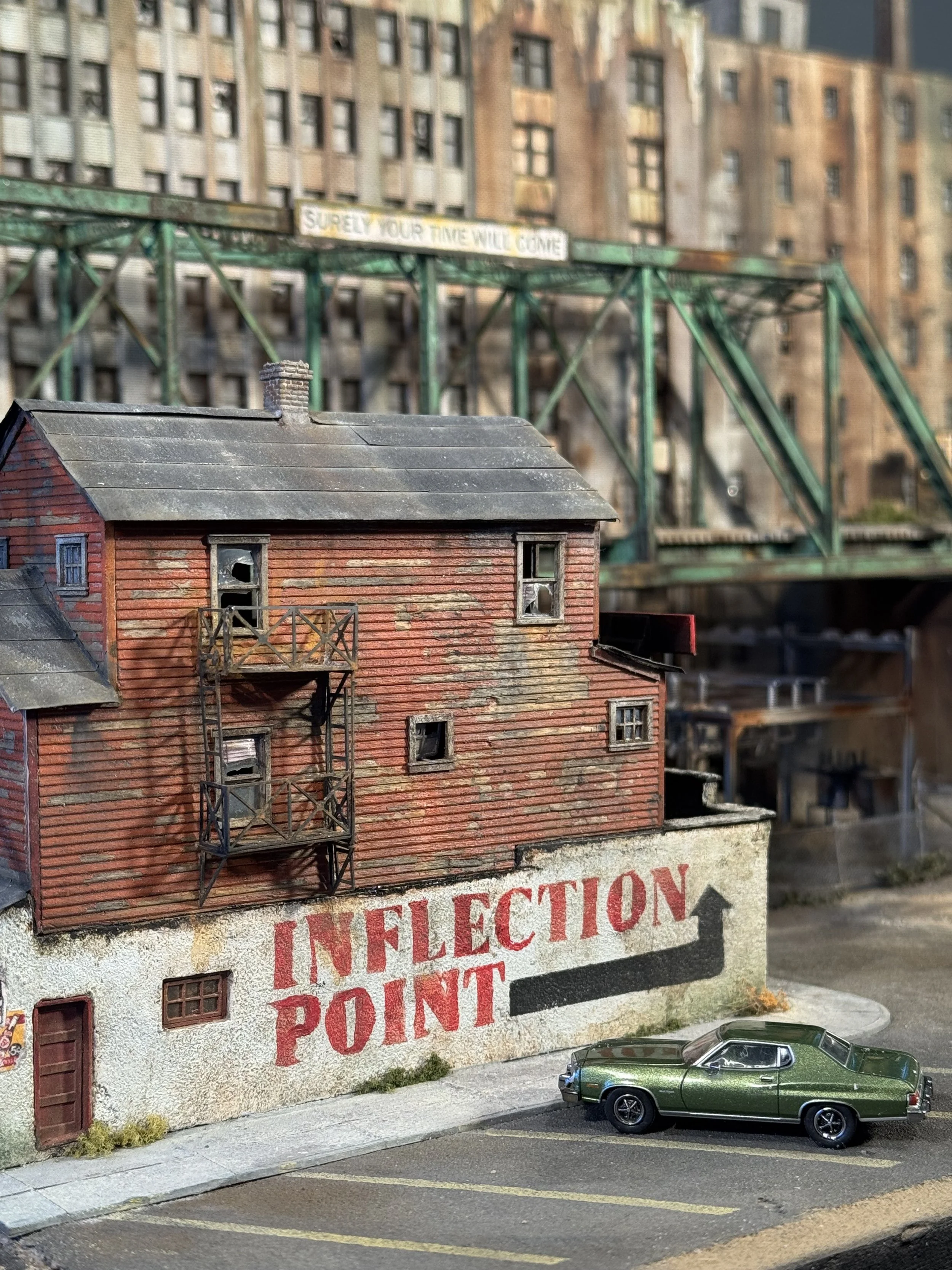 Miniature model of a brick building with multiple windows, a small balcony, and a sloped roof, situated next to a parking lot with a green classic car and a sign in the background that reads 'INFECTION POINT' and a billboard on a bridge in the backgr