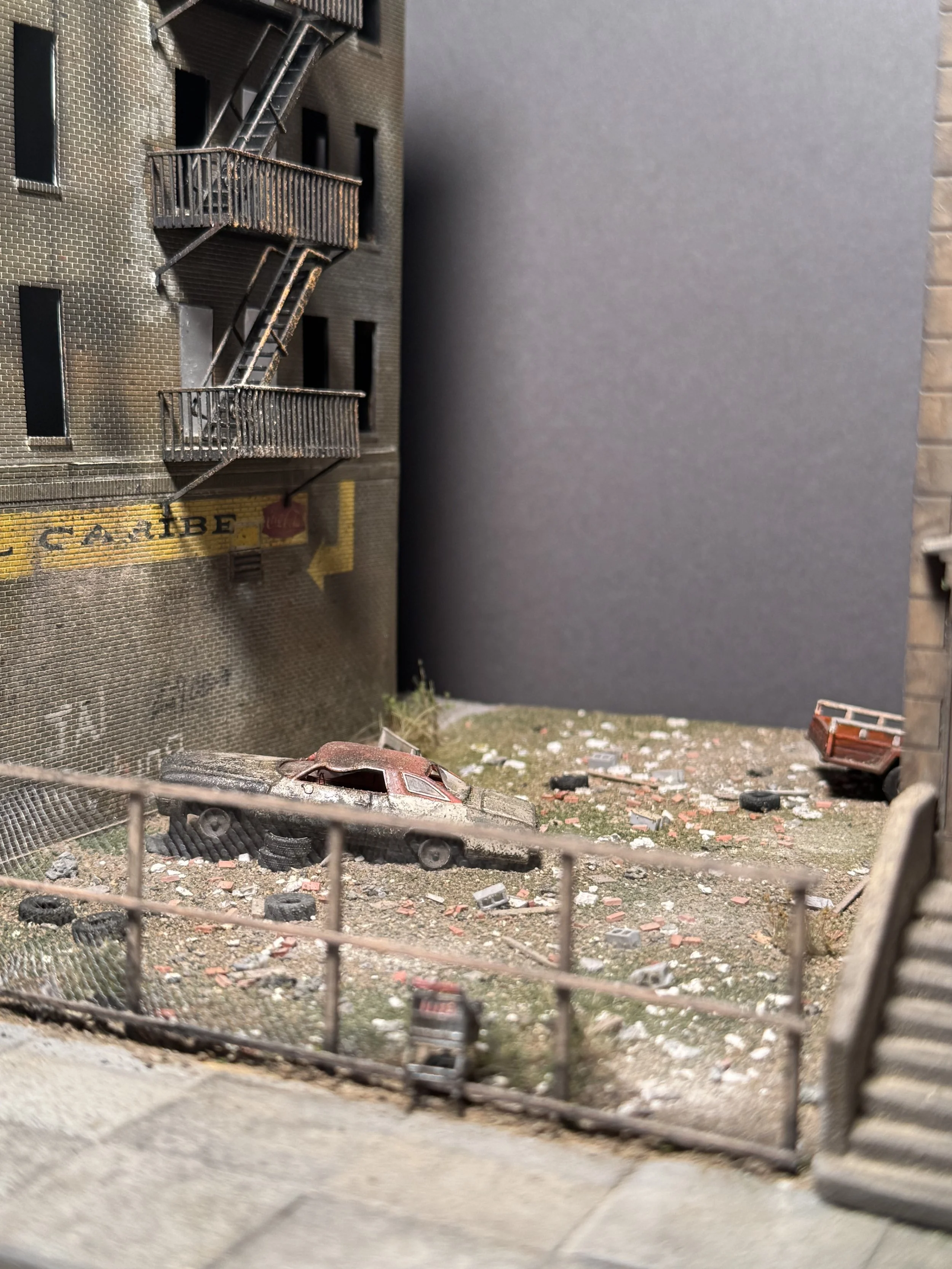 A detailed miniature scene of an urban alleyway featuring a weathered, overturned red car surrounded by scattered debris, tires, and trash, with a brick building displaying fire escapes and a partially visible painted sign on the wall.