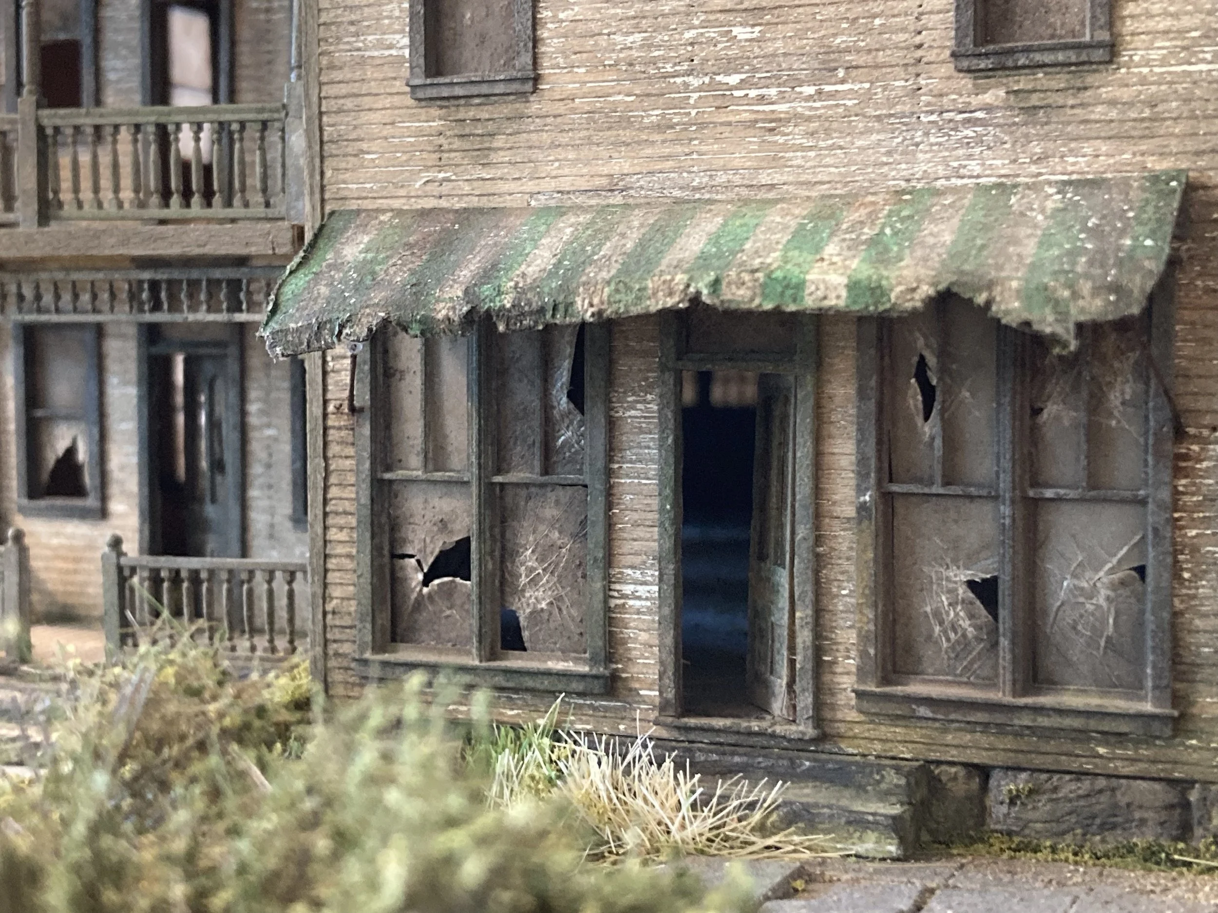 A weathered wooden porch with a striped green and brown awning, attached to an old brick house with multiple windows, some boarded up, and surrounded by green bushes.