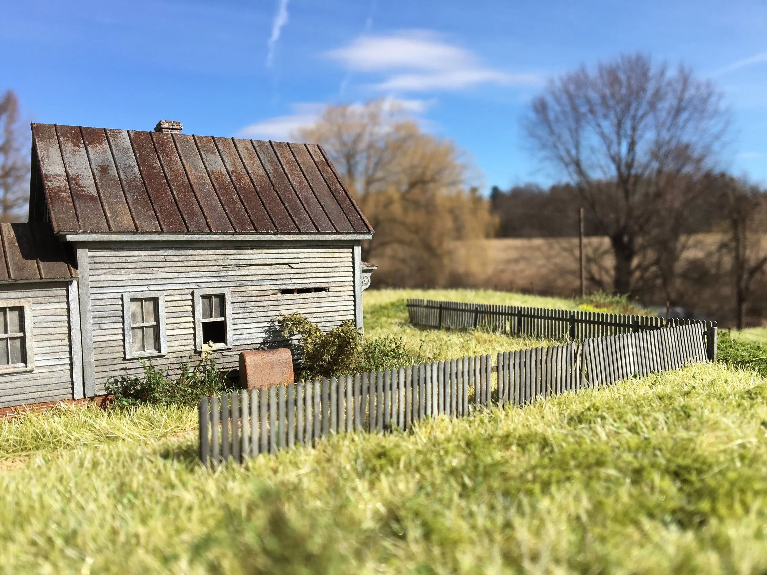 A model of an old wooden house with a rusted metal roof and small windows, surrounded by a grassy yard and a wooden fence, with leafless trees and a blue sky with clouds in the background.