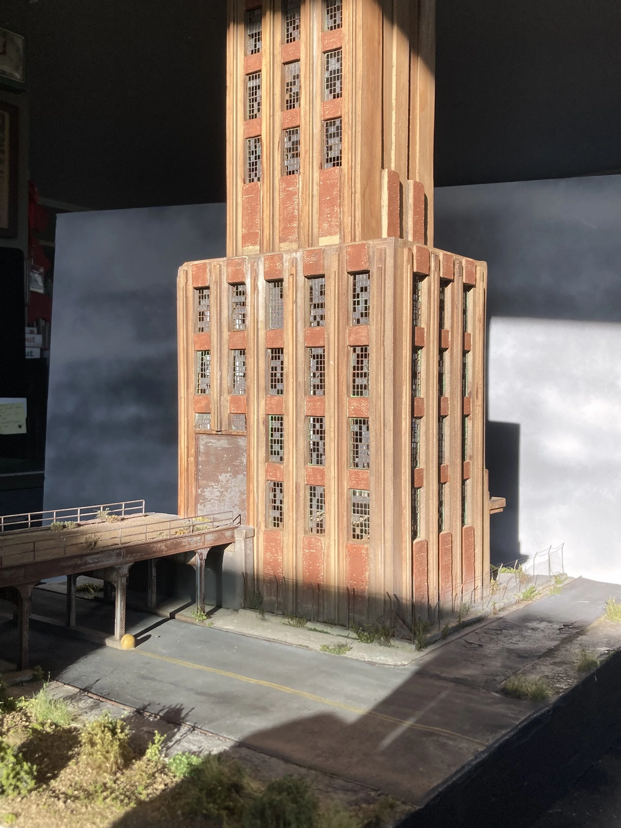 Miniature model of a tall red brick and wood building with multiple windows, set on a base with a surrounding fence and a small paved area.