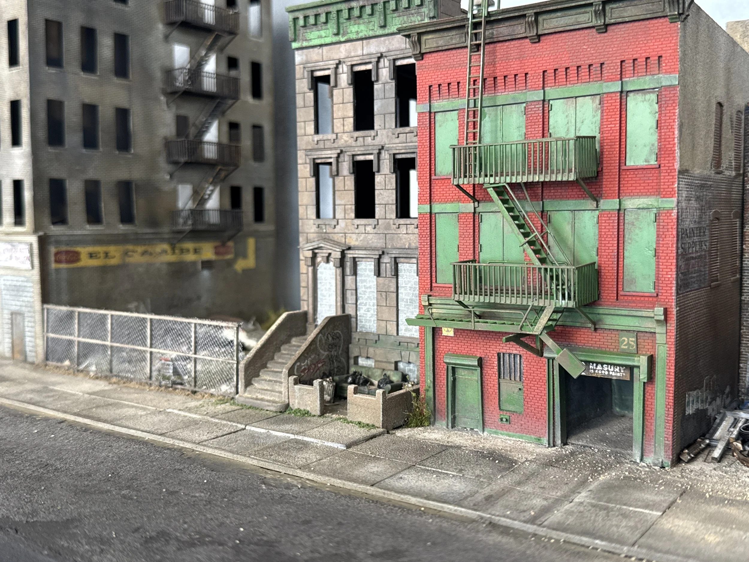 A street scene showing a red brick building with green painted fire escape stairs and balcony, adjacent to a gray building with black fire escapes, and a chain-link fence and concrete stairs leading up to the buildings.