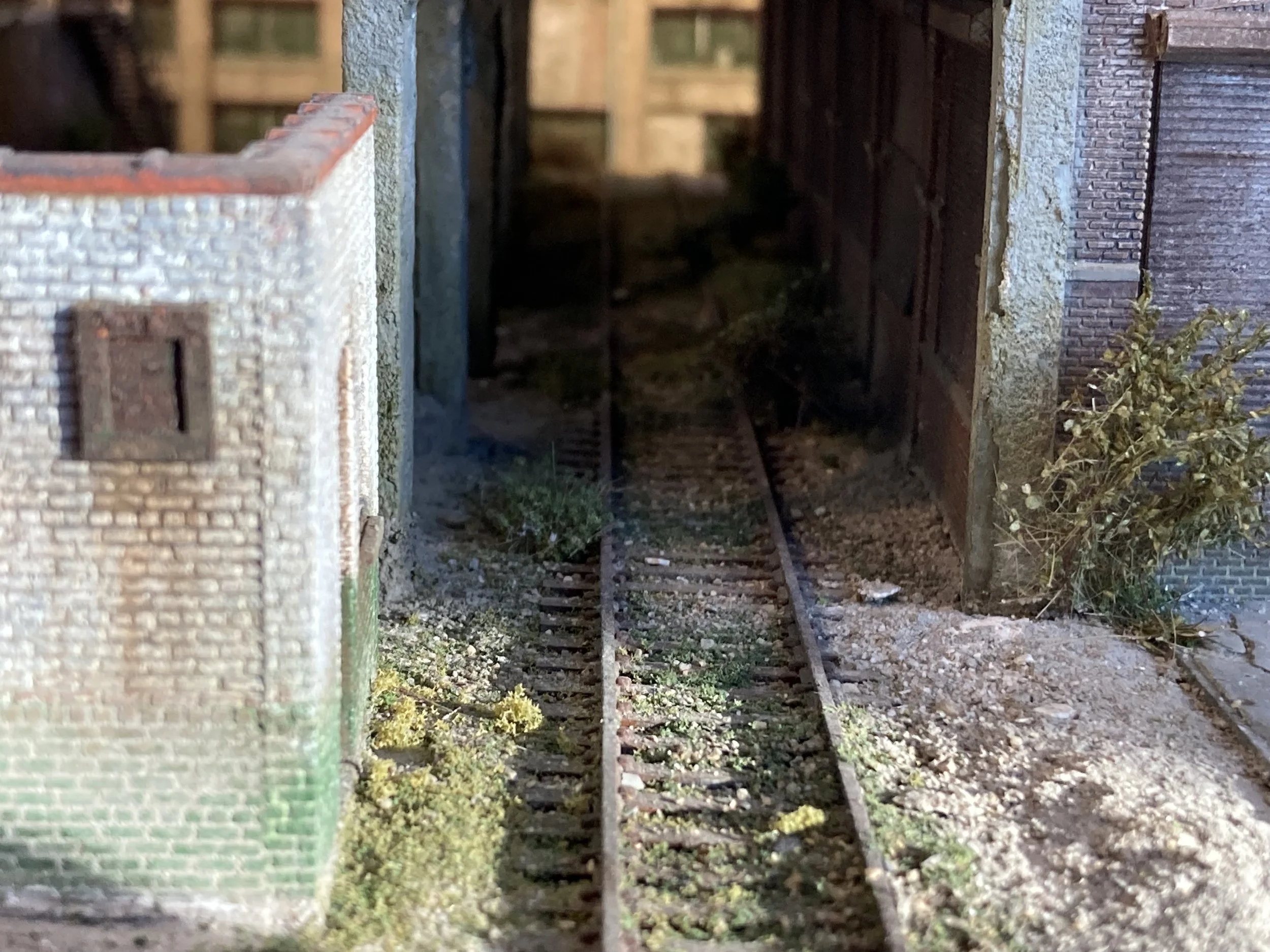 Miniature model of a old brick building, narrow alley with train tracks, small plants on the ground, and a dark passageway in the background.