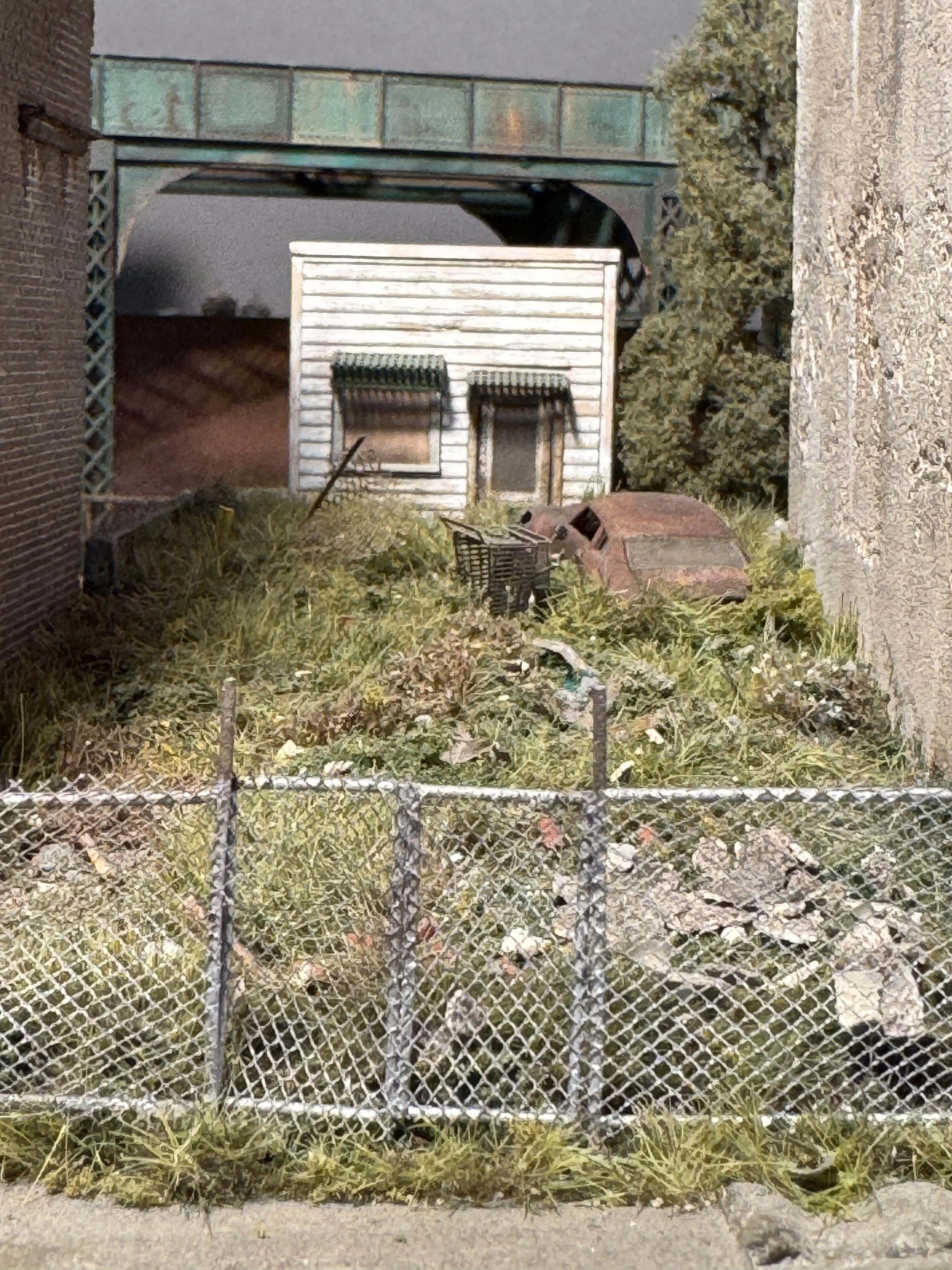 A small abandoned backyard with overgrown grass, a rusted shopping cart, a discarded brick structure, and a white shed with a window and door, enclosed by a chain-link fence.