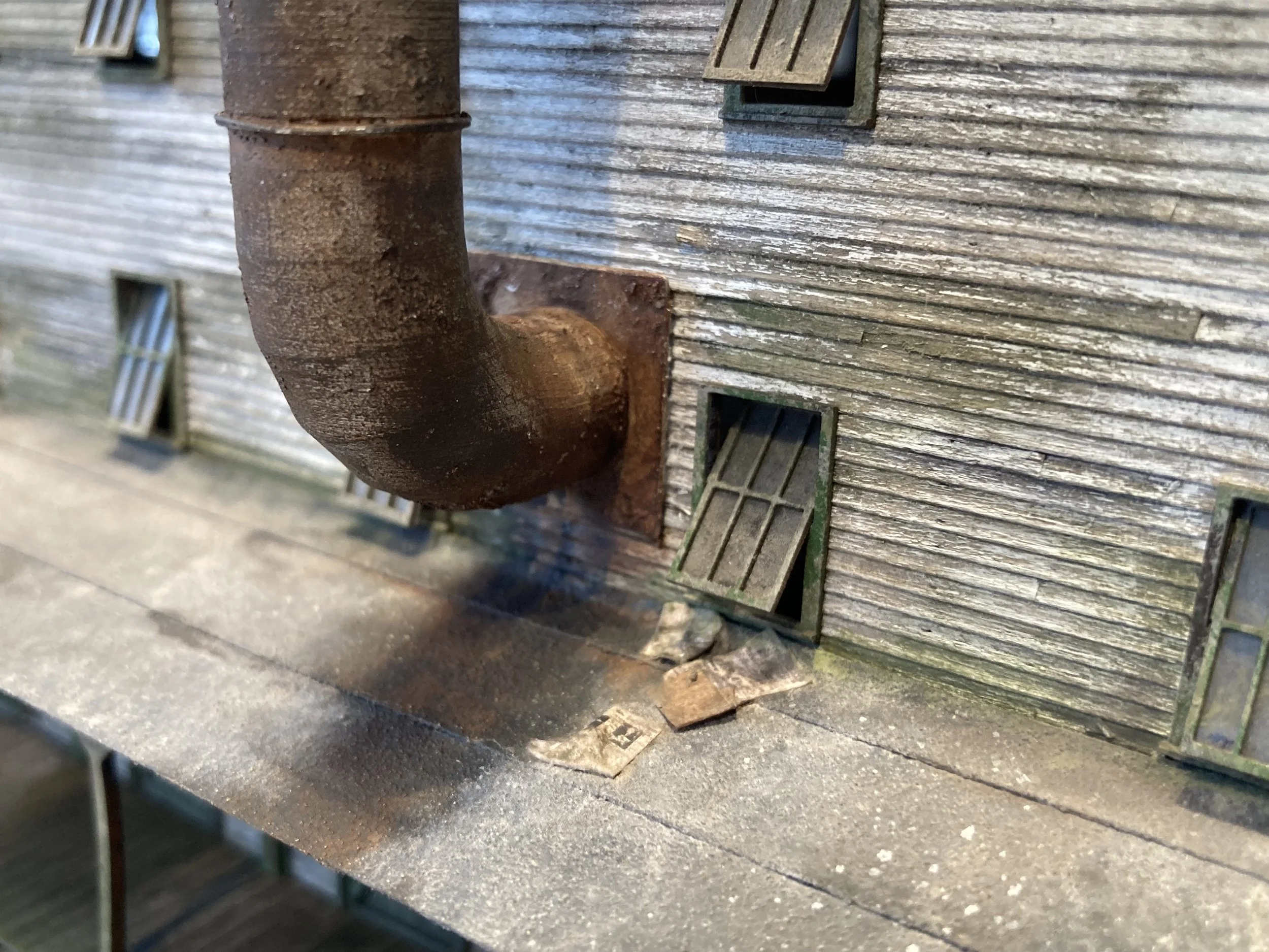 A weathered wooden exterior wall with a rusty metal pipe attached, and small square windows with slatted wooden shutters.