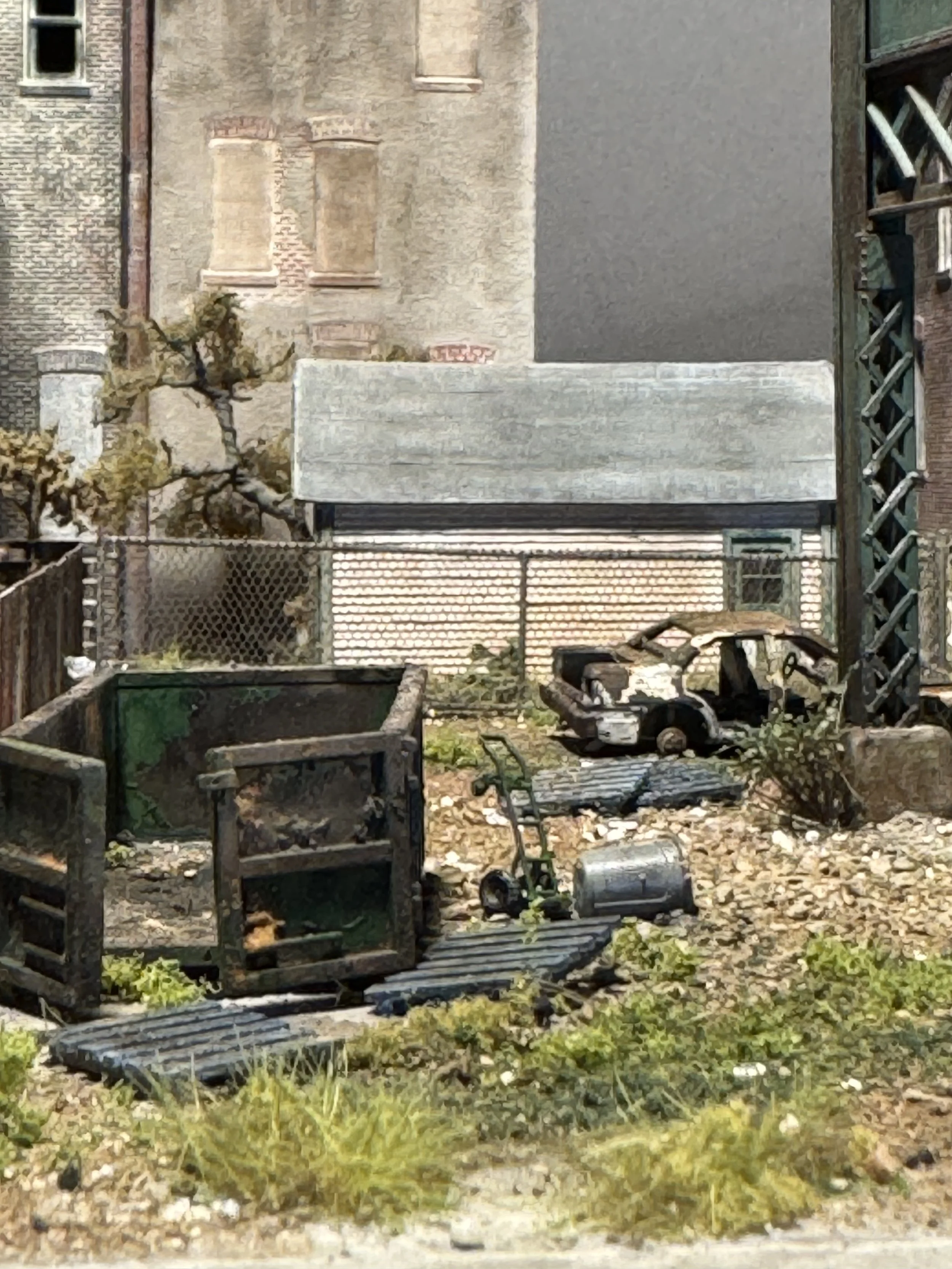 Backyard with garden tools, a wheelbarrow, and a few potted plants, enclosed with a metal fence, adjacent to old brick buildings.