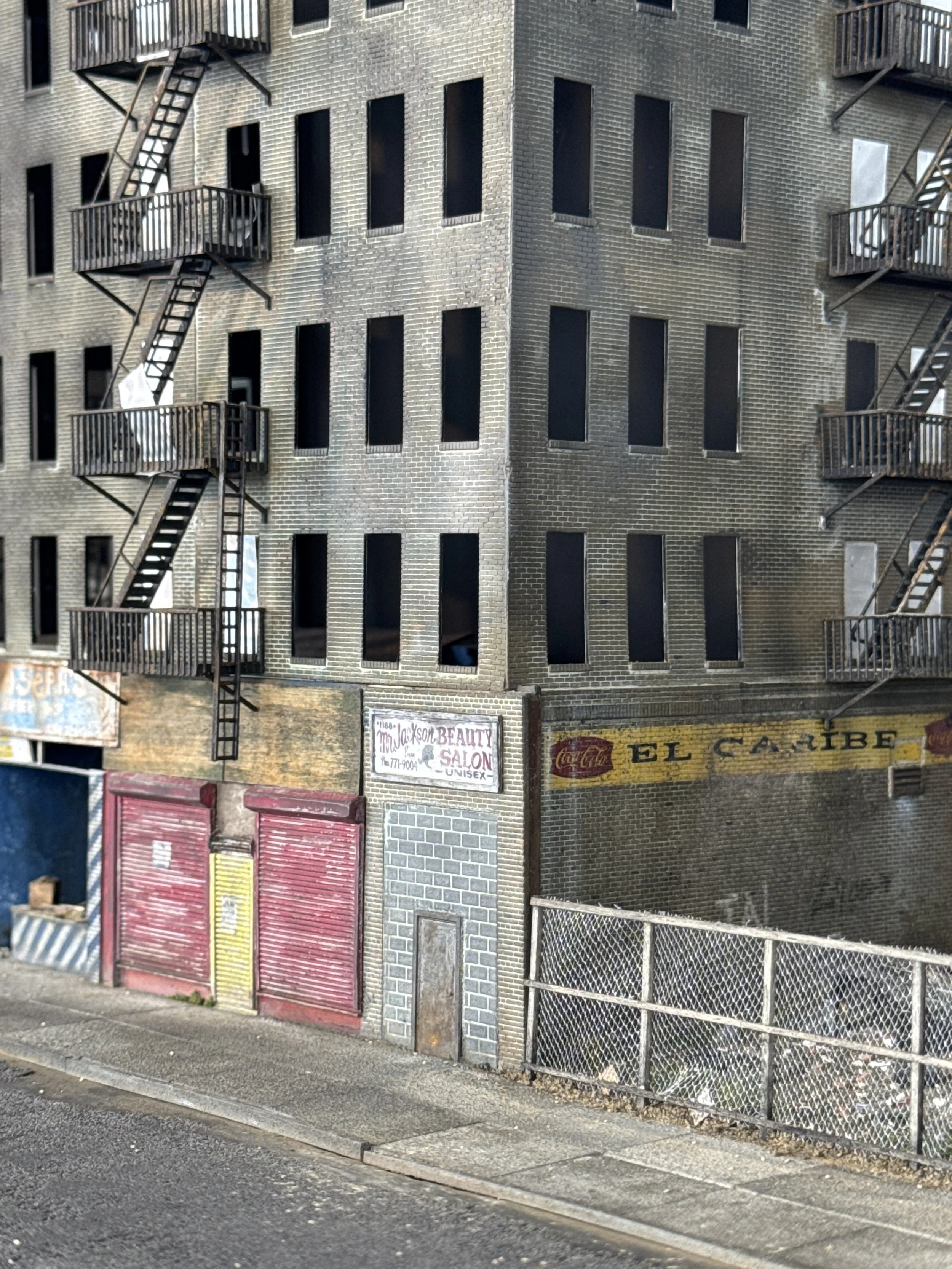 A multi-story brick building with black rectangular windows and fire escapes on the side. The ground floor has various business storefronts, including a beauty salon and a grocery store, with colorful closed shutters. There is a sidewalk in front and