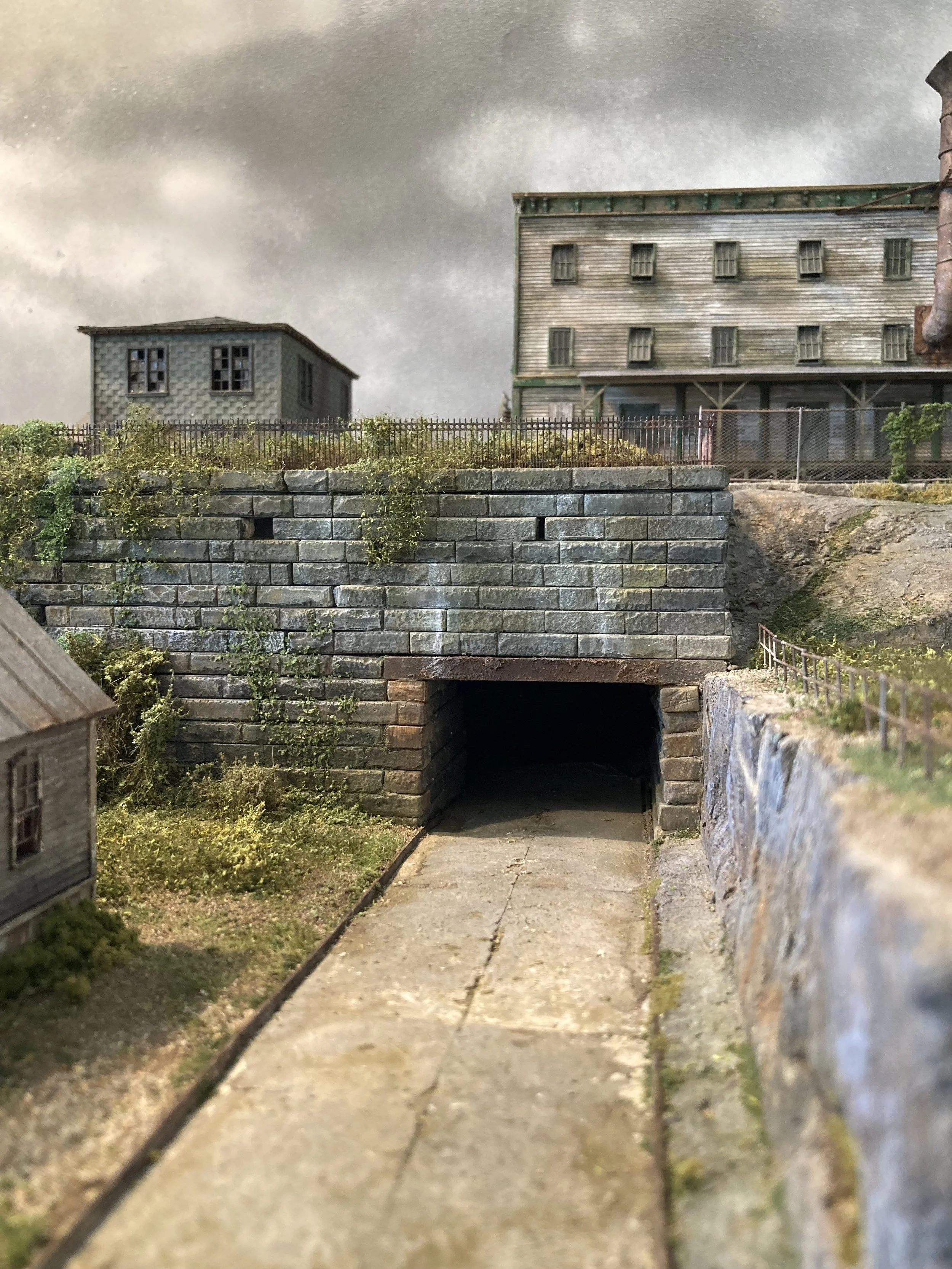 Miniature model scene of an alley with a tunnel under a stone wall, small buildings, and a cloudy sky overhead.