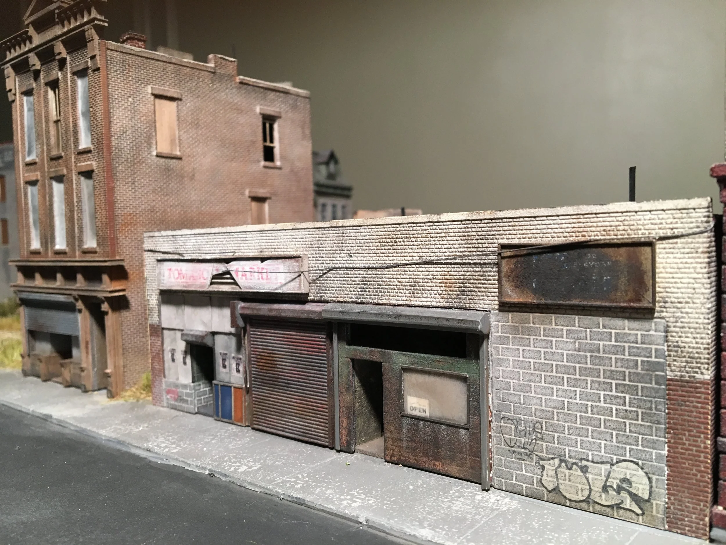 Miniature model of an urban street scene with brick buildings, a brick wall, and closed storefronts, displayed indoors.
