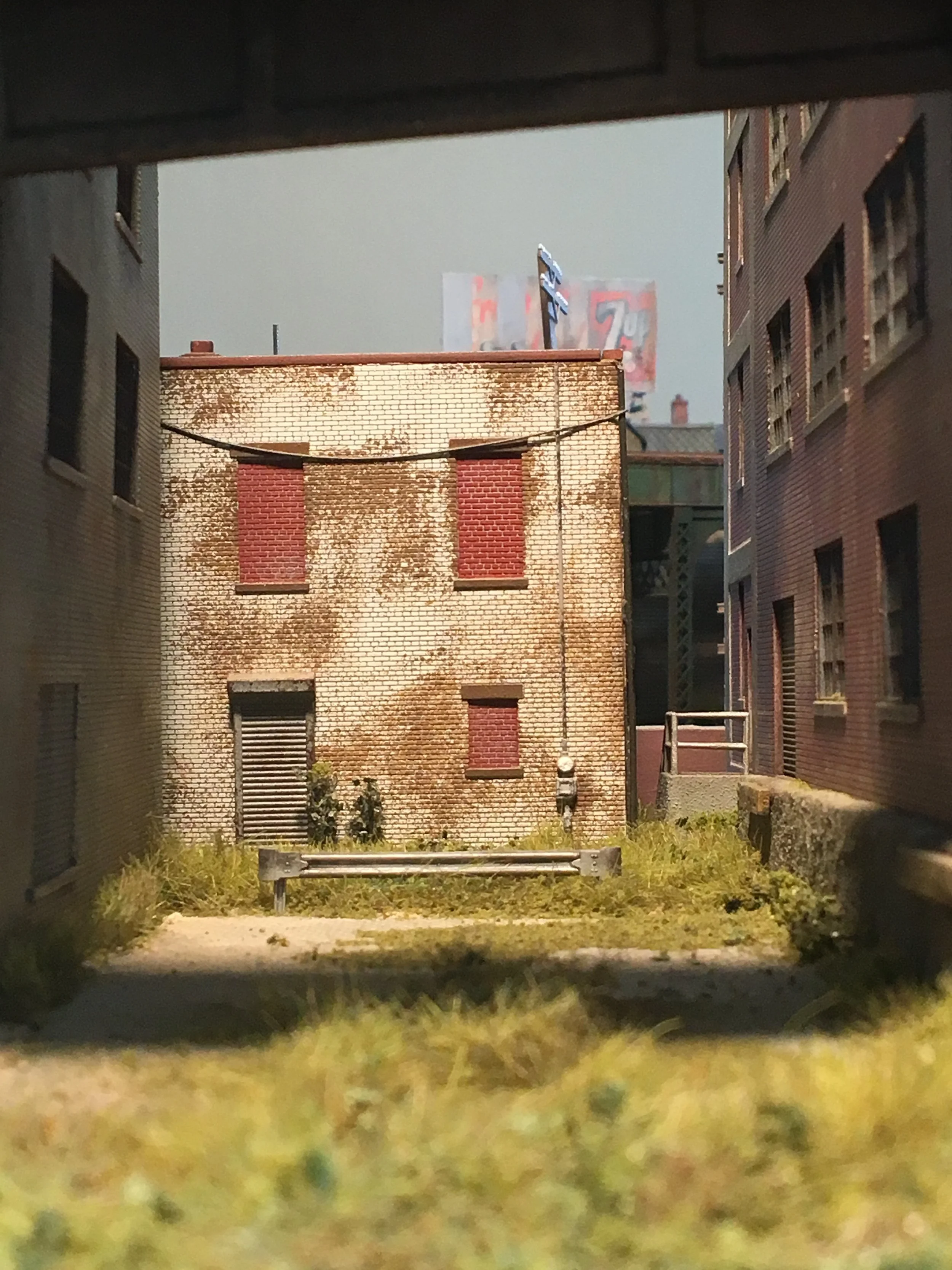 View through a small rectangular opening showing a grassy area with a bench, surrounded by brick and concrete buildings, and a billboard in the background.