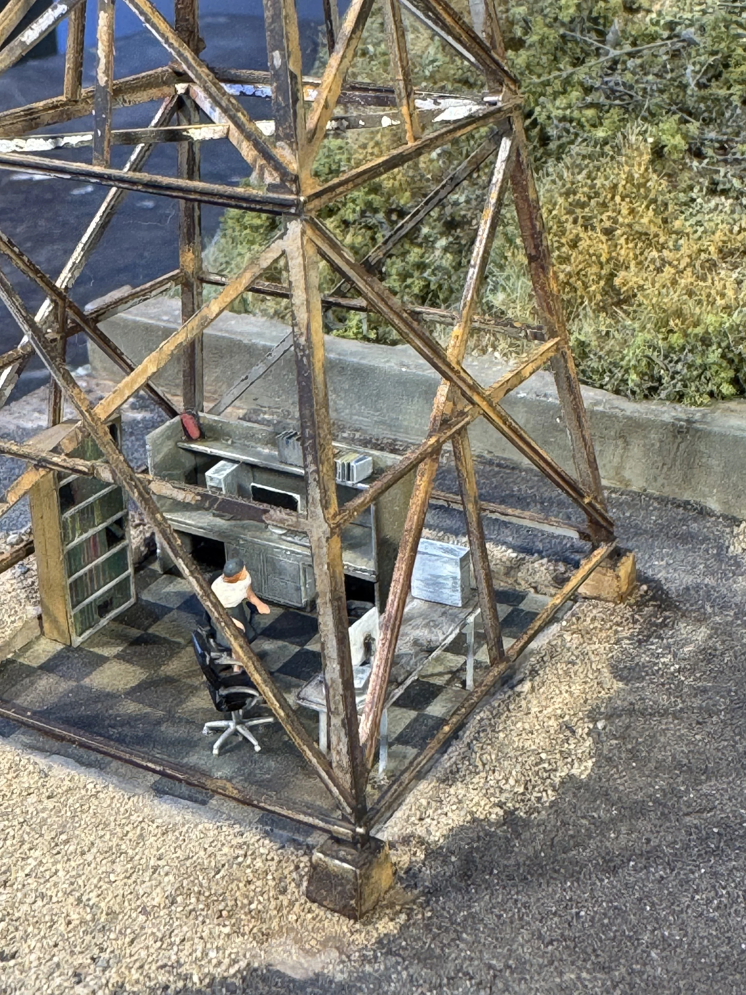 A rusty metal tower with a triangular framework viewed from below. There are electrical components and a person working at the bottom inside a small enclosed area. The ground is gravel, and there are trees with green foliage in the background.