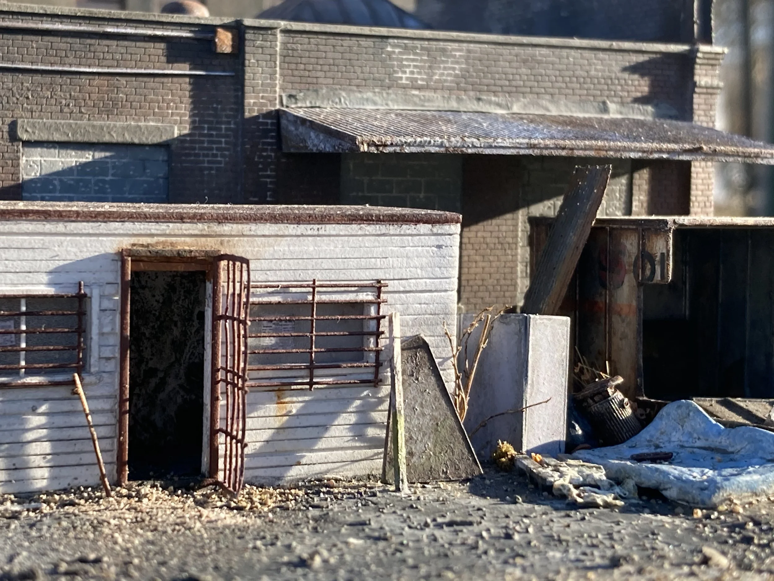 A construction site with a concrete wall, metal rebar, a rusted gate, and scattered debris, including a black trash can and plastic sheeting.