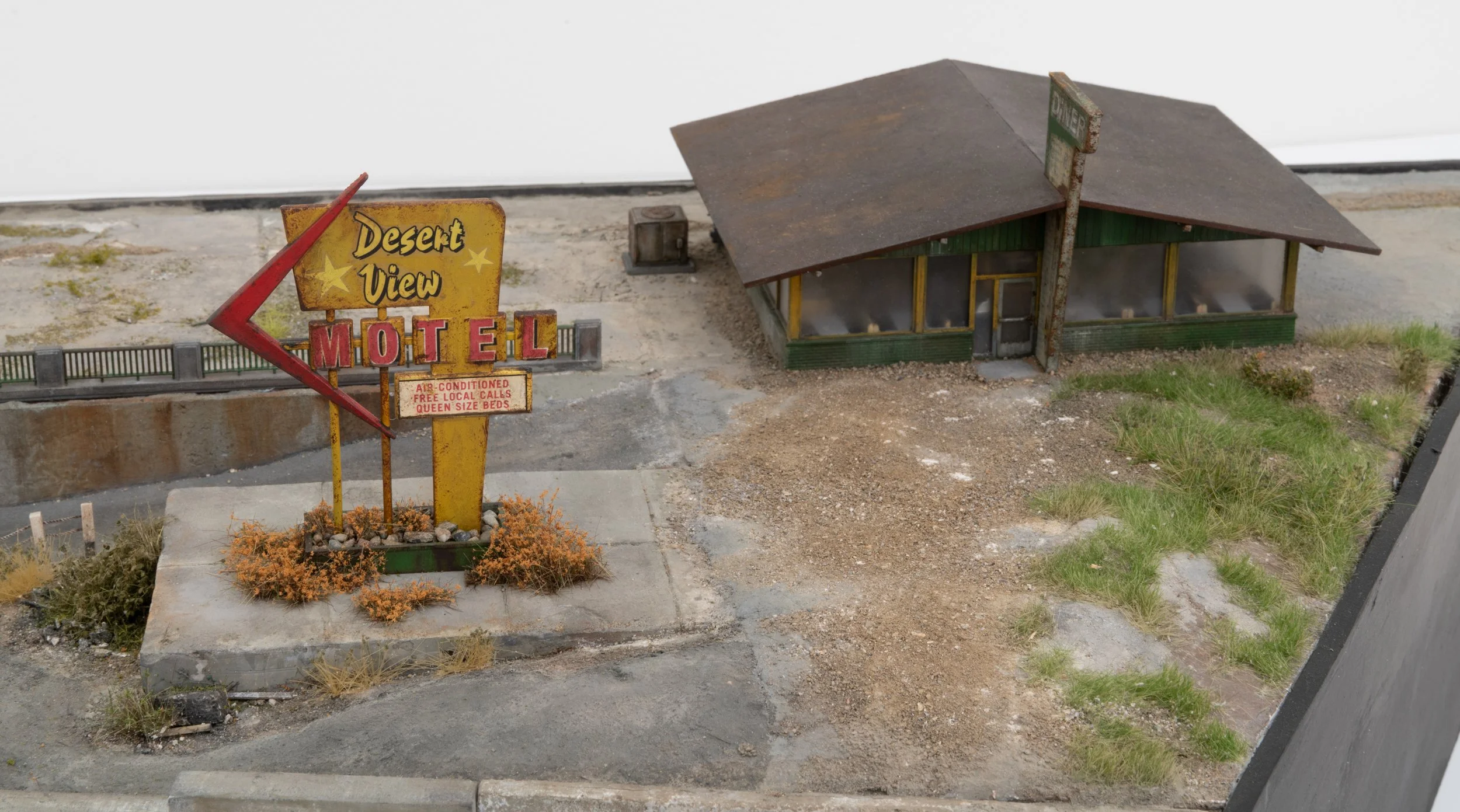 Miniature model of a building with a rusty metal roof and green walls, alongside a vintage motel sign with yellow and red lettering that reads 'Desert View Motel.' The sign features an arrow pointing left with additional text about amenities. The sur