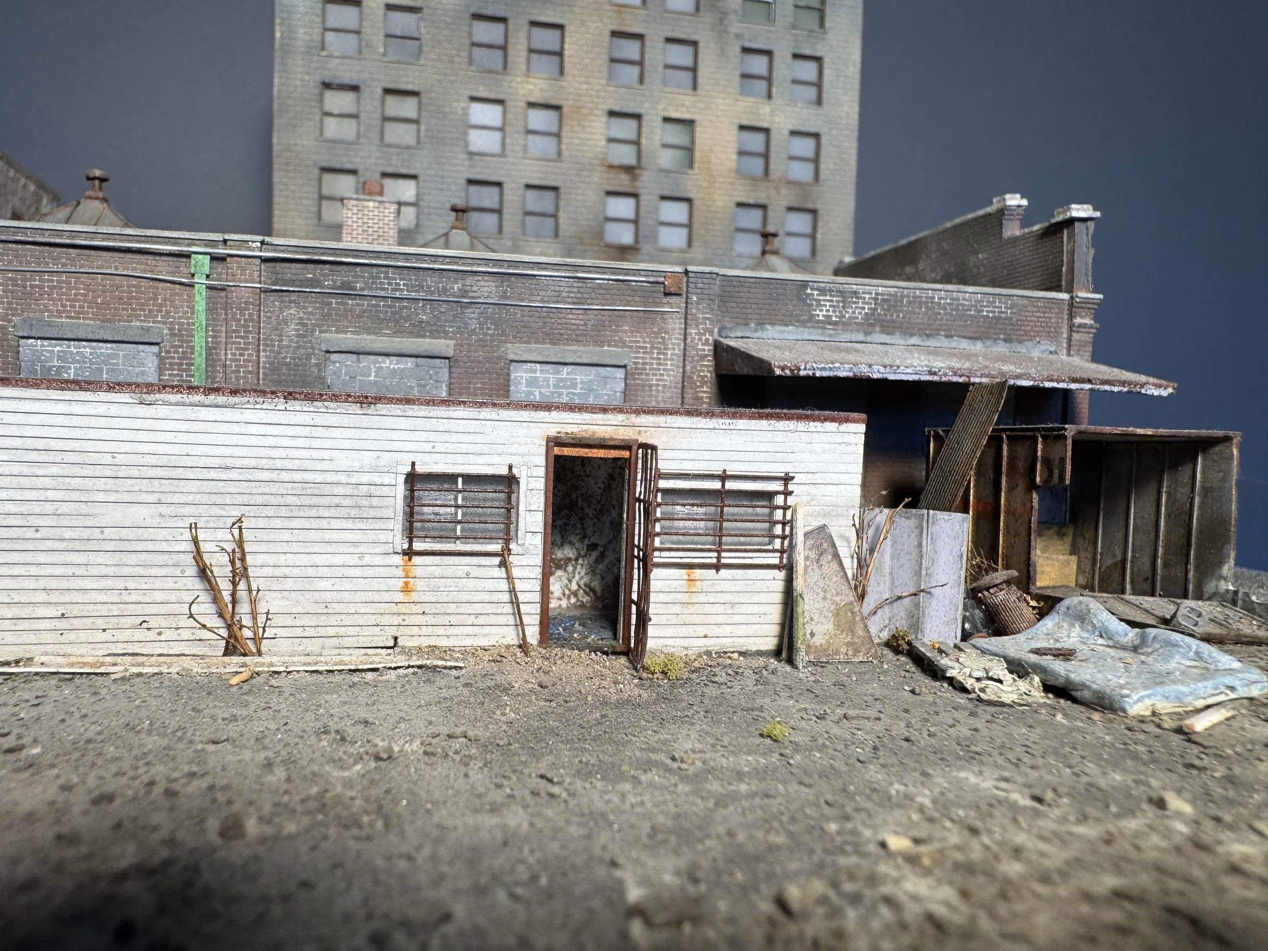 A close-up view of a dirt ground with some small debris, in front of a row of abandoned, weathered buildings with boarded-up windows and dilapidated roofing. In the background, a taller building with multiple windows is visible under a dark sky.