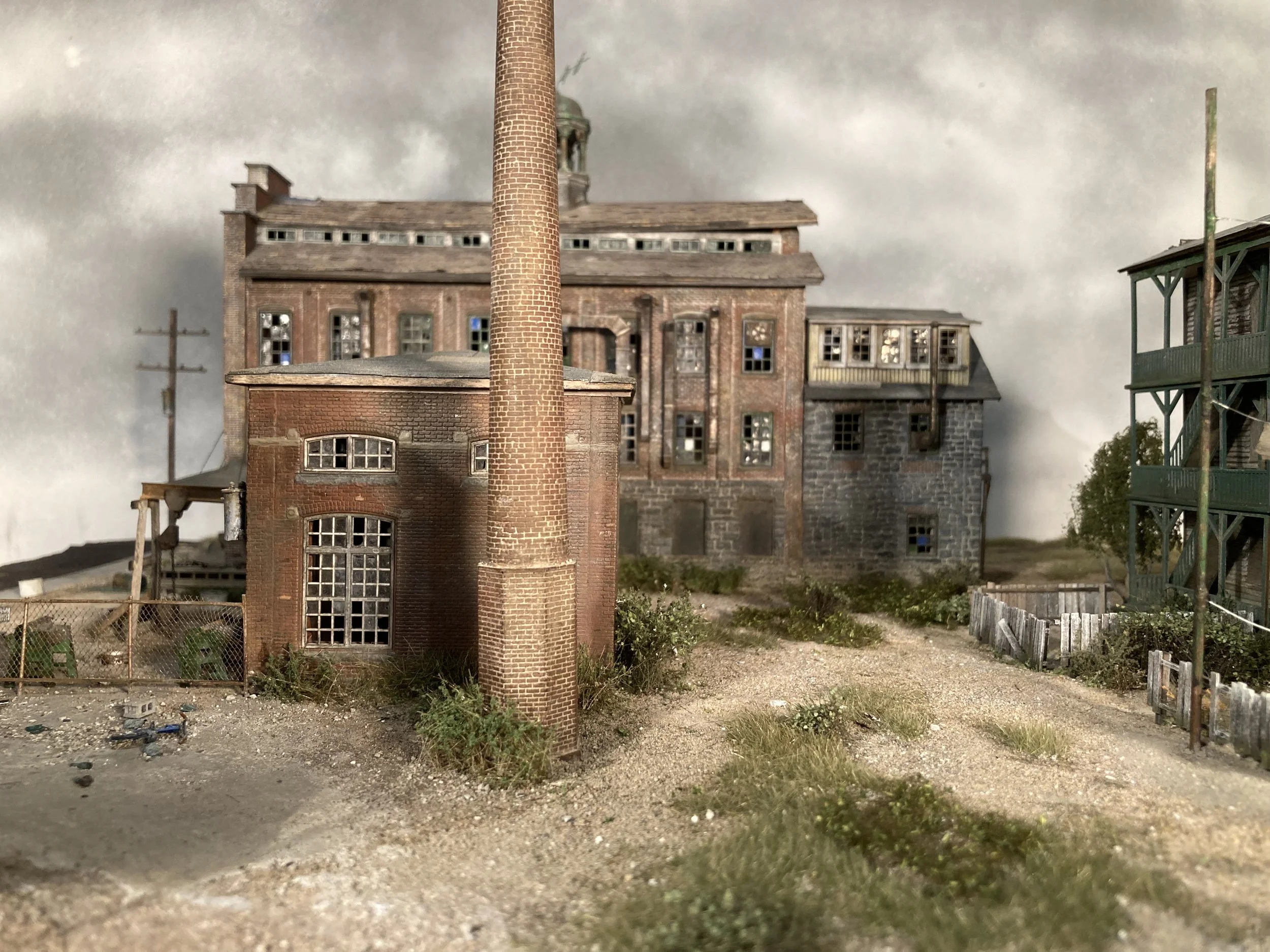 A miniature model of an old industrial building with a tall chimney, surrounded by a dirt ground, a wooden fence, and some scattered debris.
