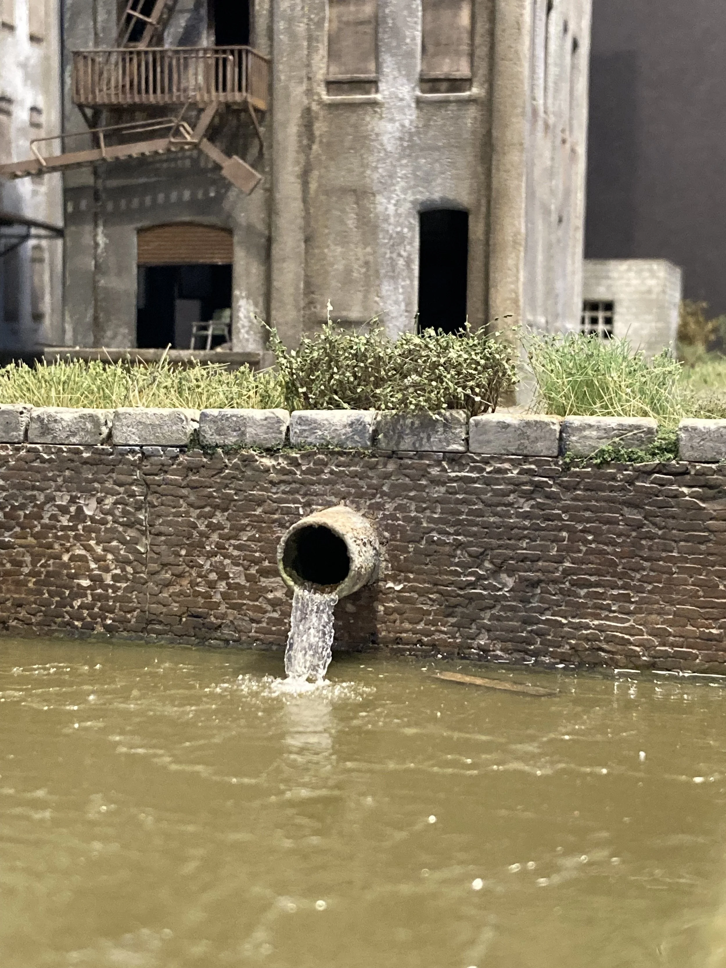 An old brick wall with a pipe spout from which water flows into a muddy river. Behind the wall, an abandoned building with boarded-up windows, a fire escape, and worn walls is visible.