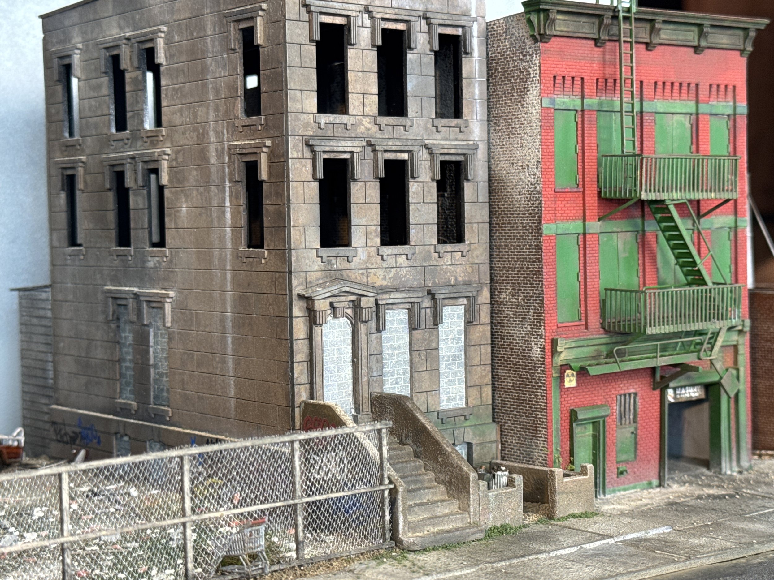Two model buildings with different architectural styles, surrounded by a metal fence. The building on the left is a grey stone-style structure with multiple window frames and decorative details. The building on the right is a red brick style with gre
