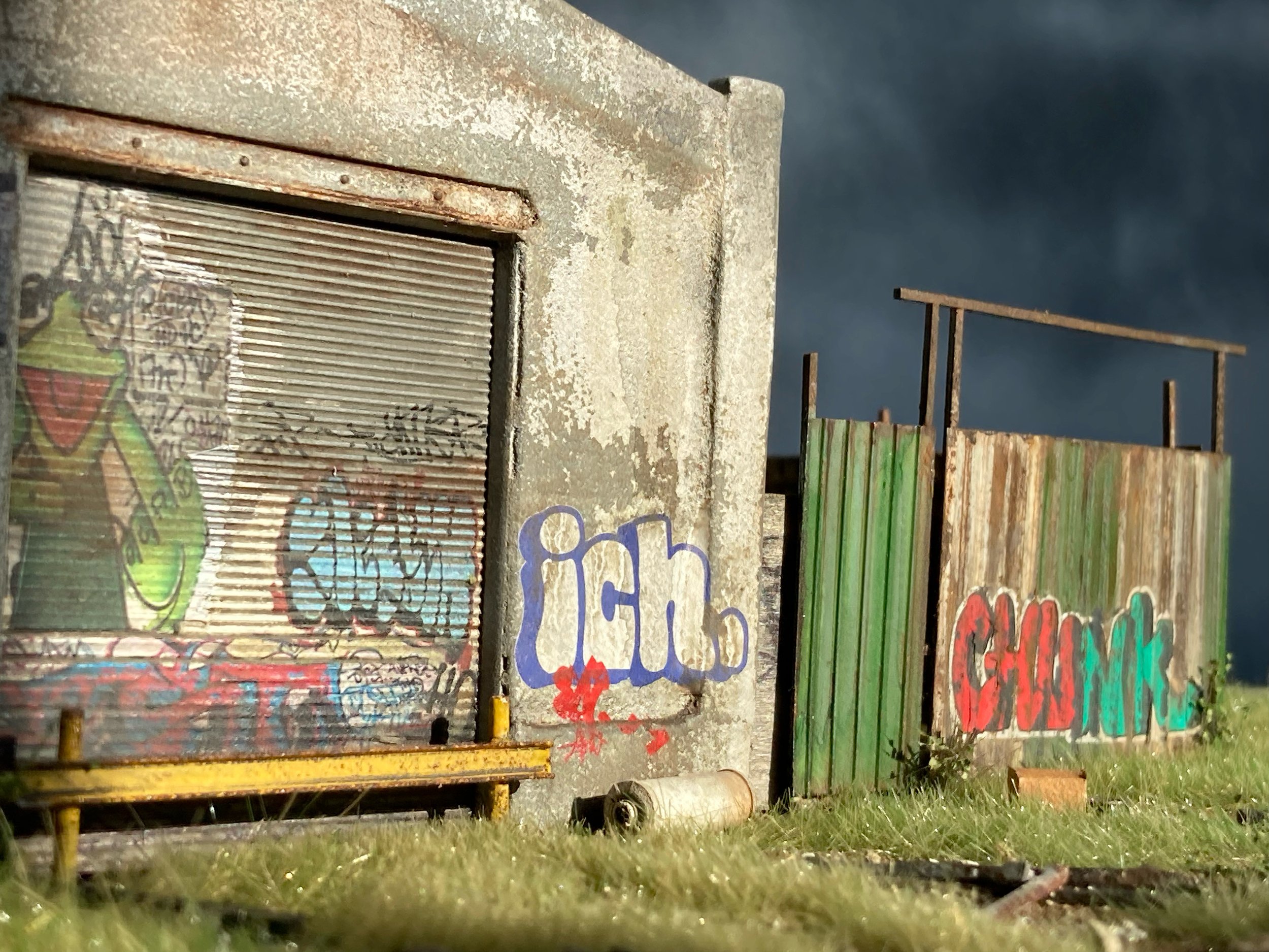 A rusty, weathered metal structure with graffiti and a small cardboard sign, situated outdoors on grass, under a dark sky.