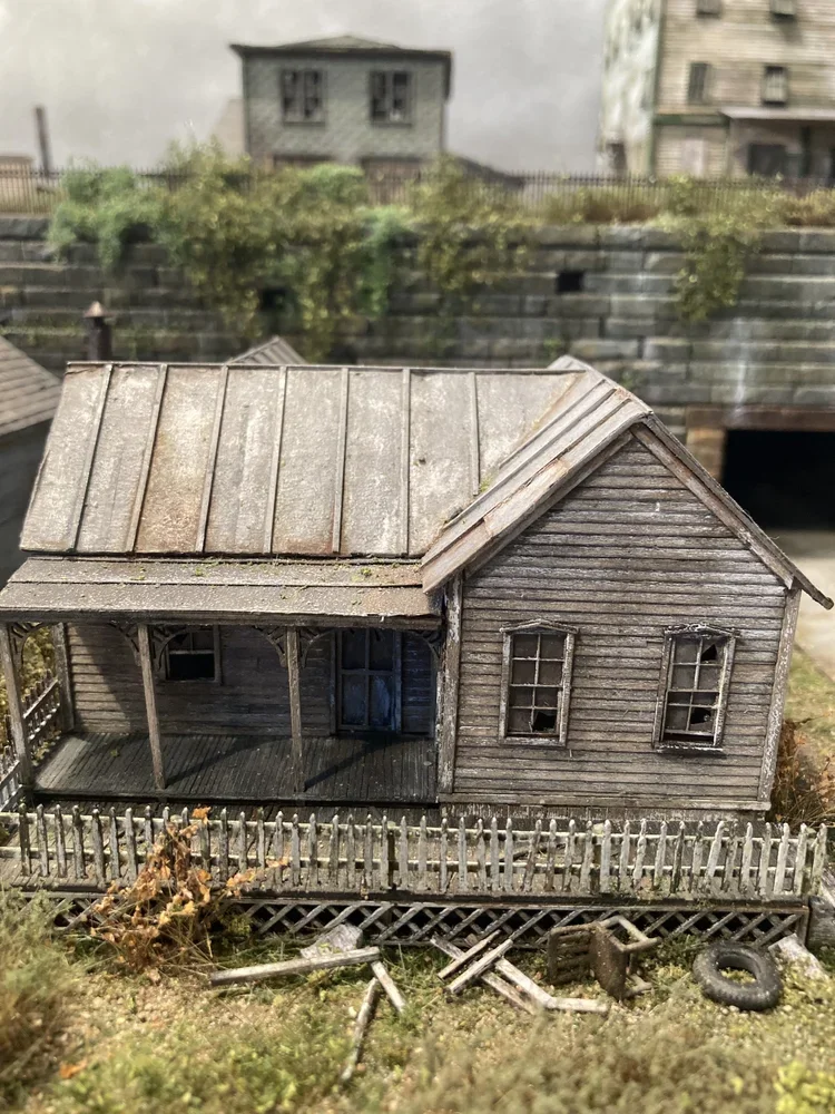 Miniature model of an old, weathered house with a porch and broken window, surrounded by a small yard with debris and a tire, in a detailed diorama setting.