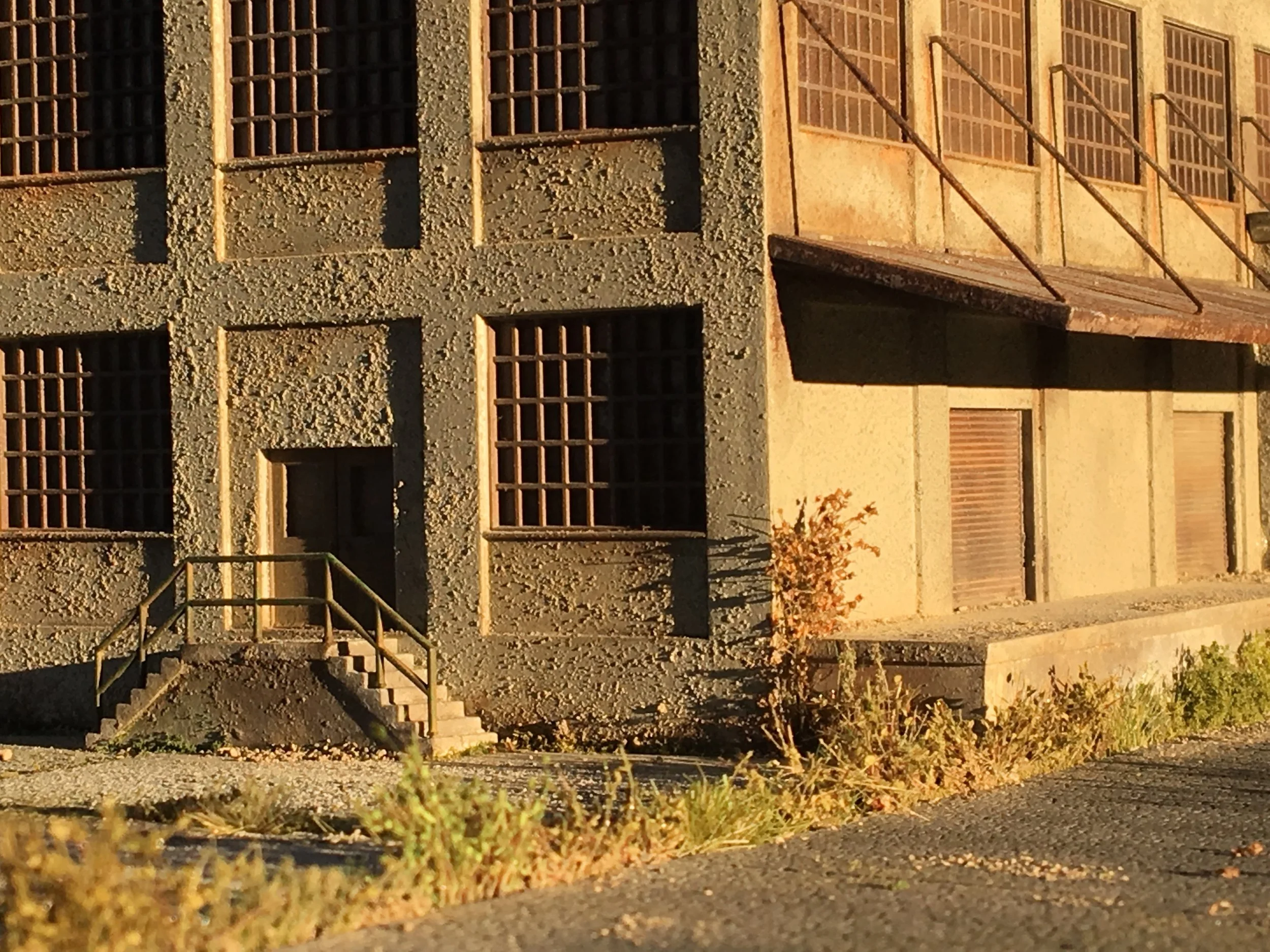 A view of an industrial building with a textured concrete exterior, barred windows, and a small staircase outside. The building appears to be old, with staircase railings and shadows visible in the sunlight.