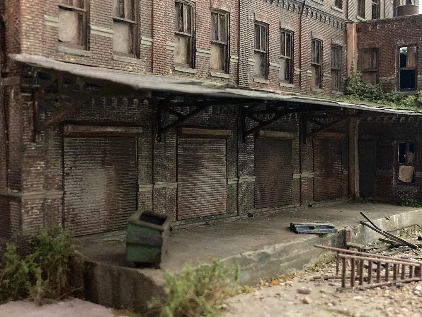 An abandoned brick building with boarded-up windows and closed metal shutters, some greenery and debris on the sidewalk.
