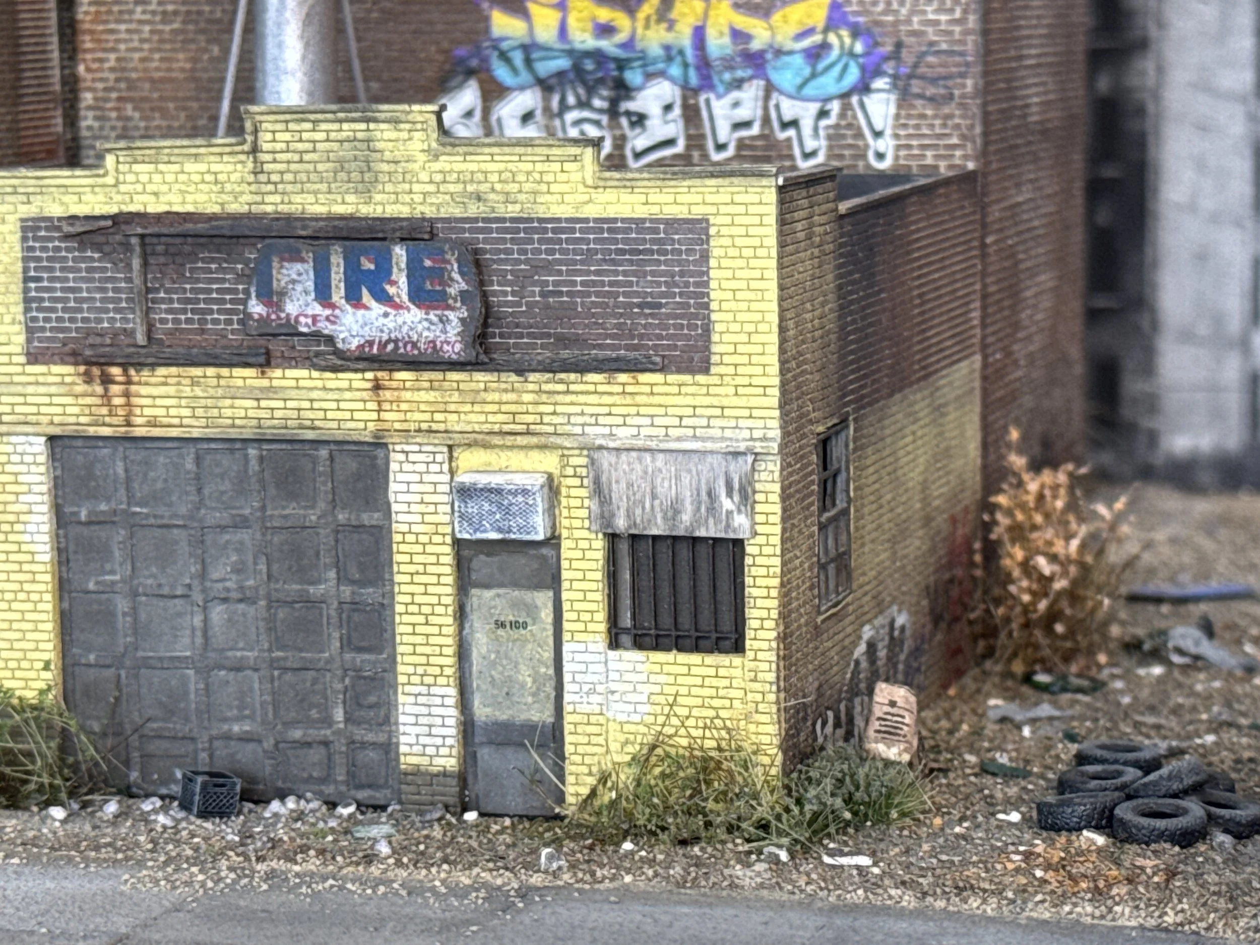 A miniature model of an urban building with yellow brick walls and a garage door at the bottom. There is a faded, weathered sign that says 'CLIRE' on the wall above the garage. The surroundings include gravel, some debris, and sparse vegetation.