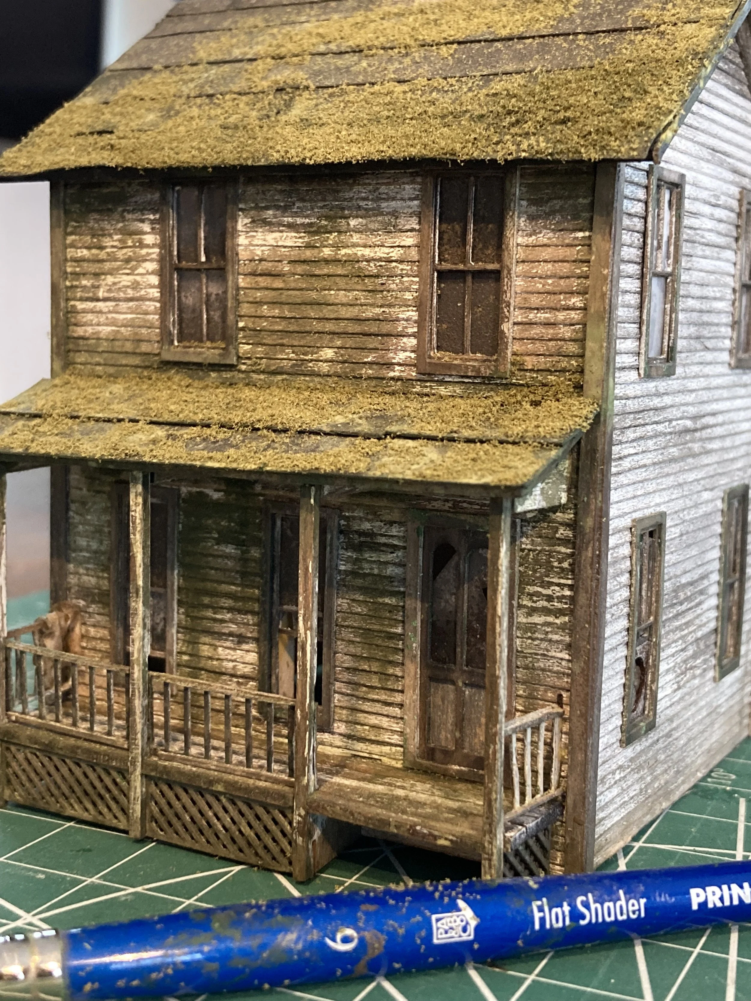 A miniature model of a two-story wooden house with a porch, set on a green cutting mat with a blue marker in the foreground.
