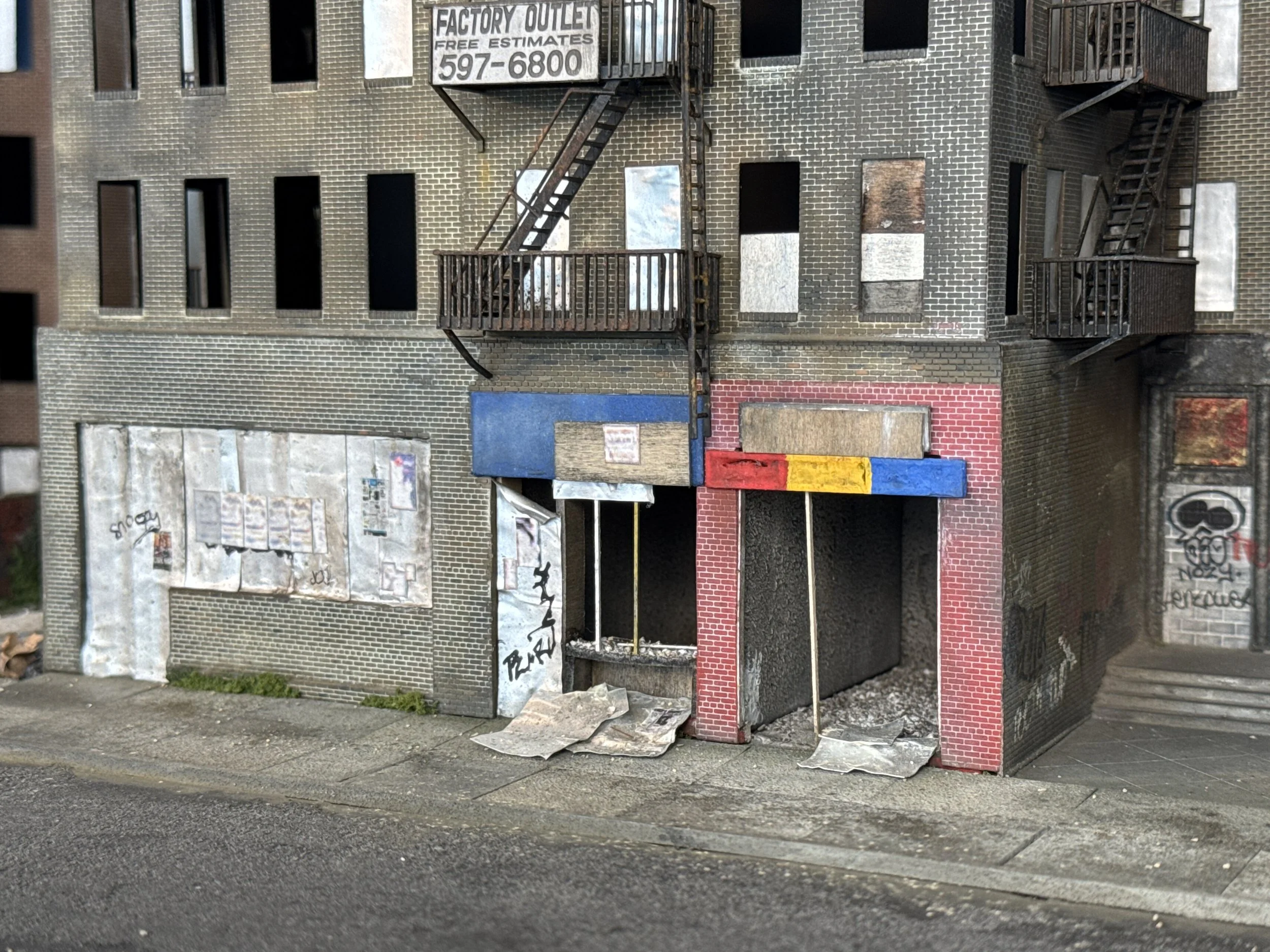 A multi-story brick building with fire escape stairs, boarded-up windows, and graffiti on the walls, located next to a parking lot and sidewalk.