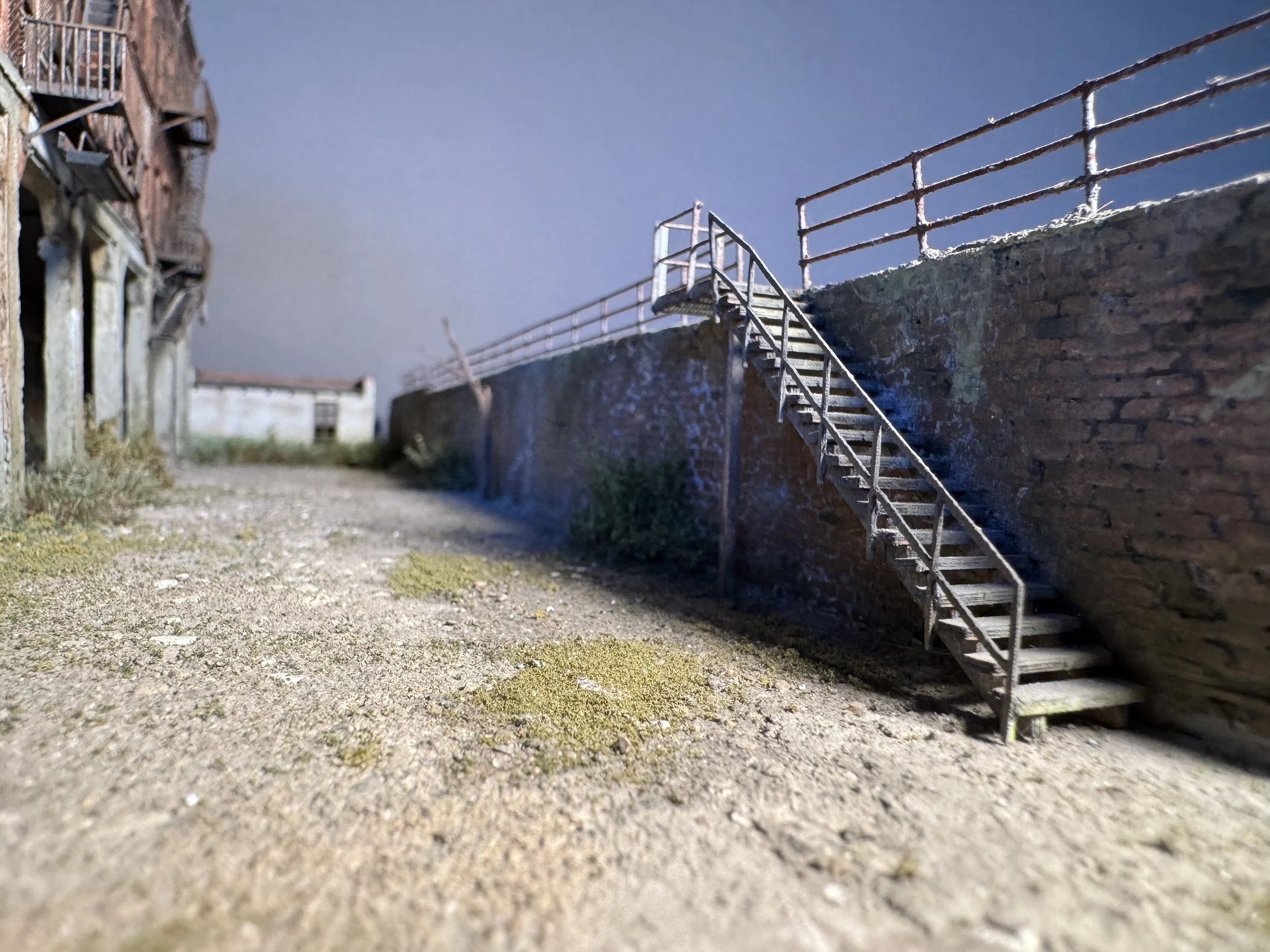 A small outdoor scene with old, weathered buildings, a brick wall with a staircase, and a gravel ground.