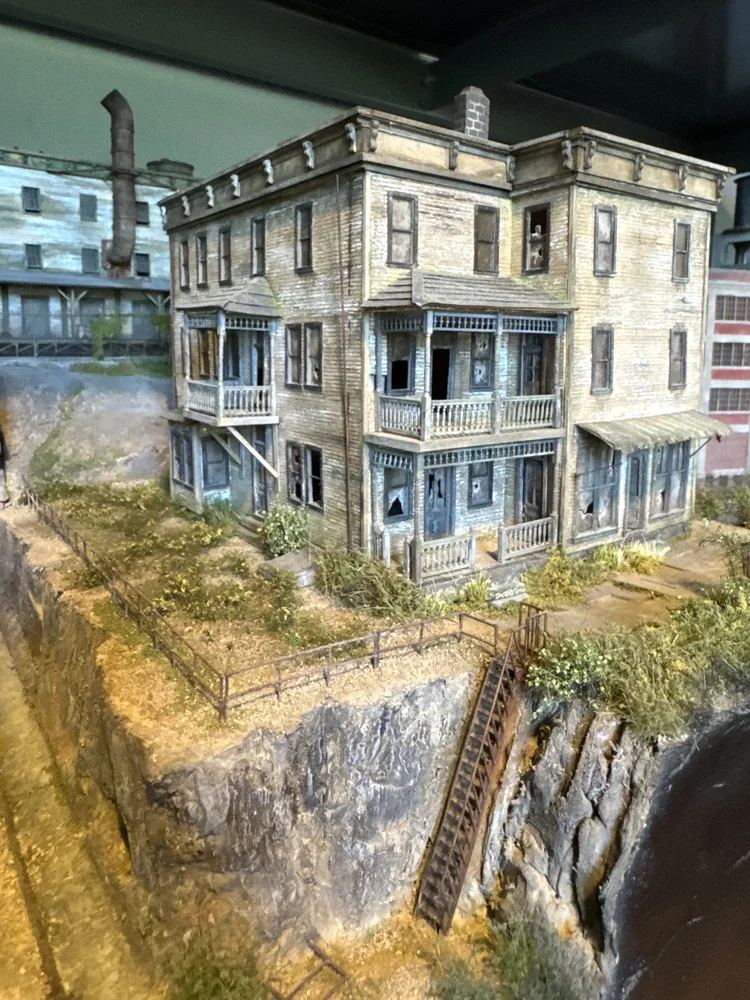 Miniature model of an old, weathered, multi-story wooden house with balconies, set on a rocky terrain with overgrown plants and a broken staircase.