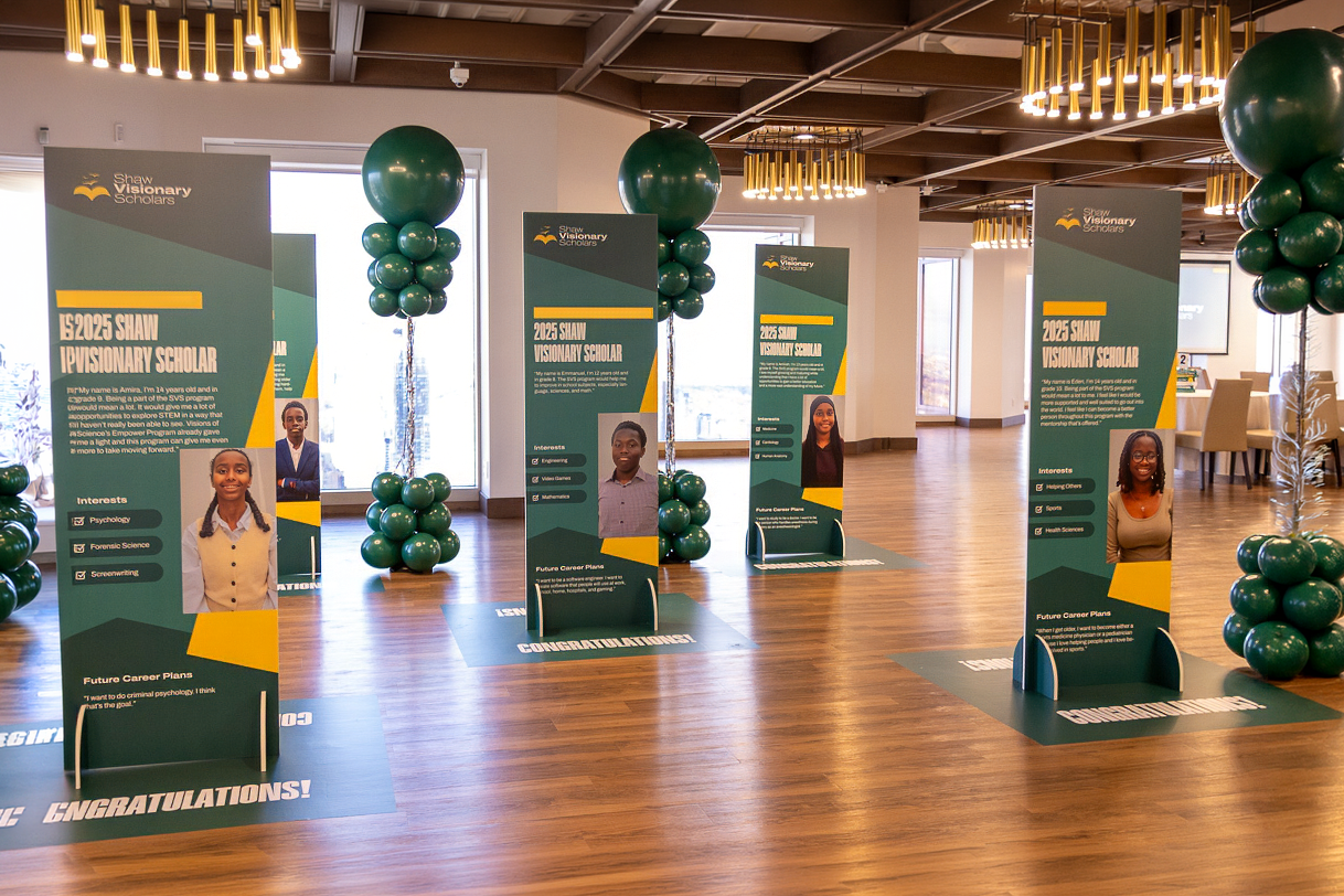 Indoor event space with green balloons and display boards featuring students' photos and information, celebrating Shaw Visionary Scholars class of 2023 and 2025.