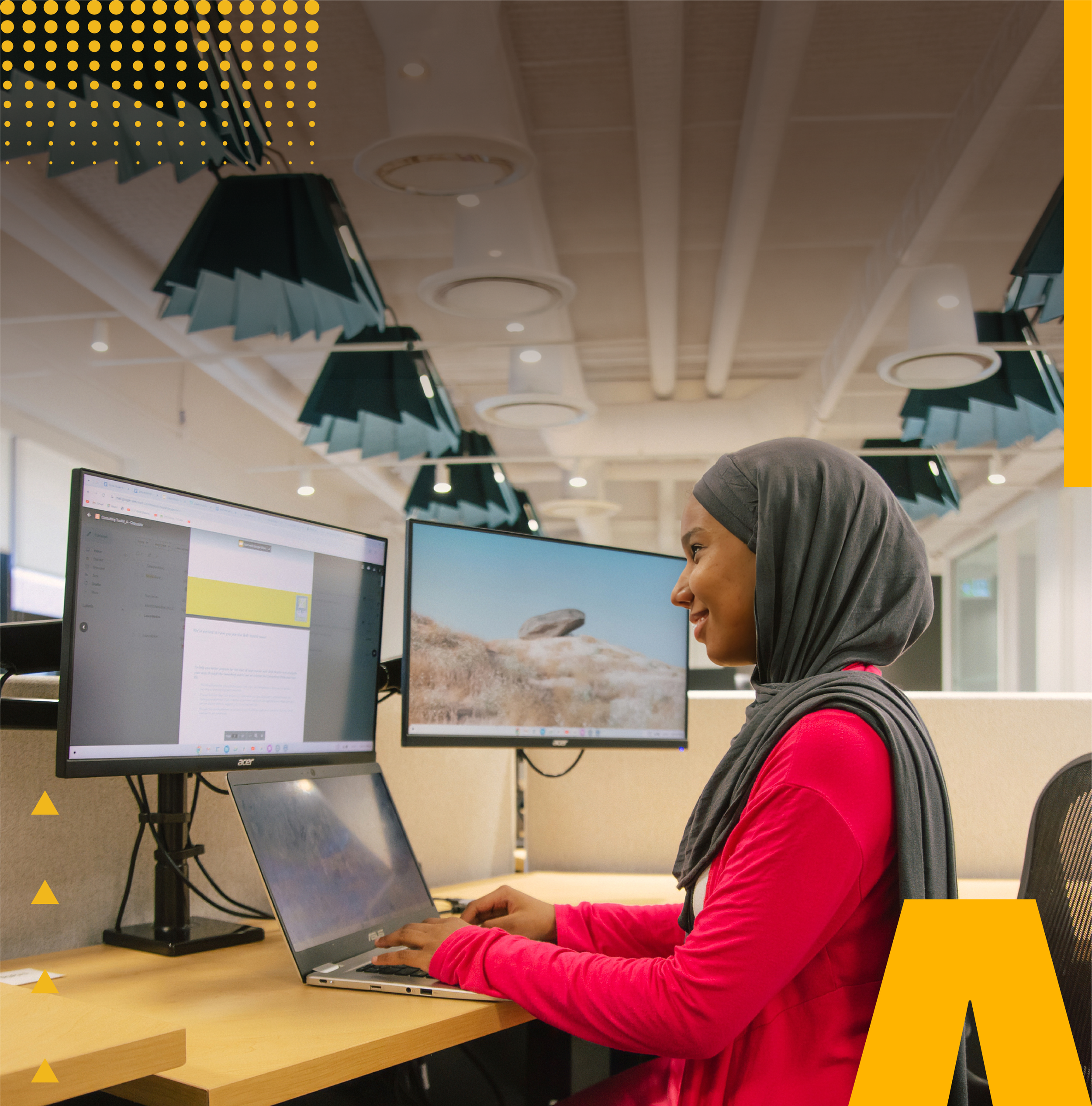 A smiling youth scholar wearing a hijab working on a laptop at a modern dual-monitor office desk for the Shaw Visionary Scholars application process.