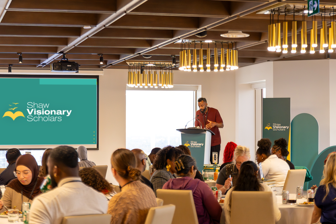 An indoor conference or seminar room with a speaker at a podium in front of an audience. The room features modern lighting fixtures, a large screen displaying the Shaw Visionary Scholars logo, and attendees sitting at tables.
