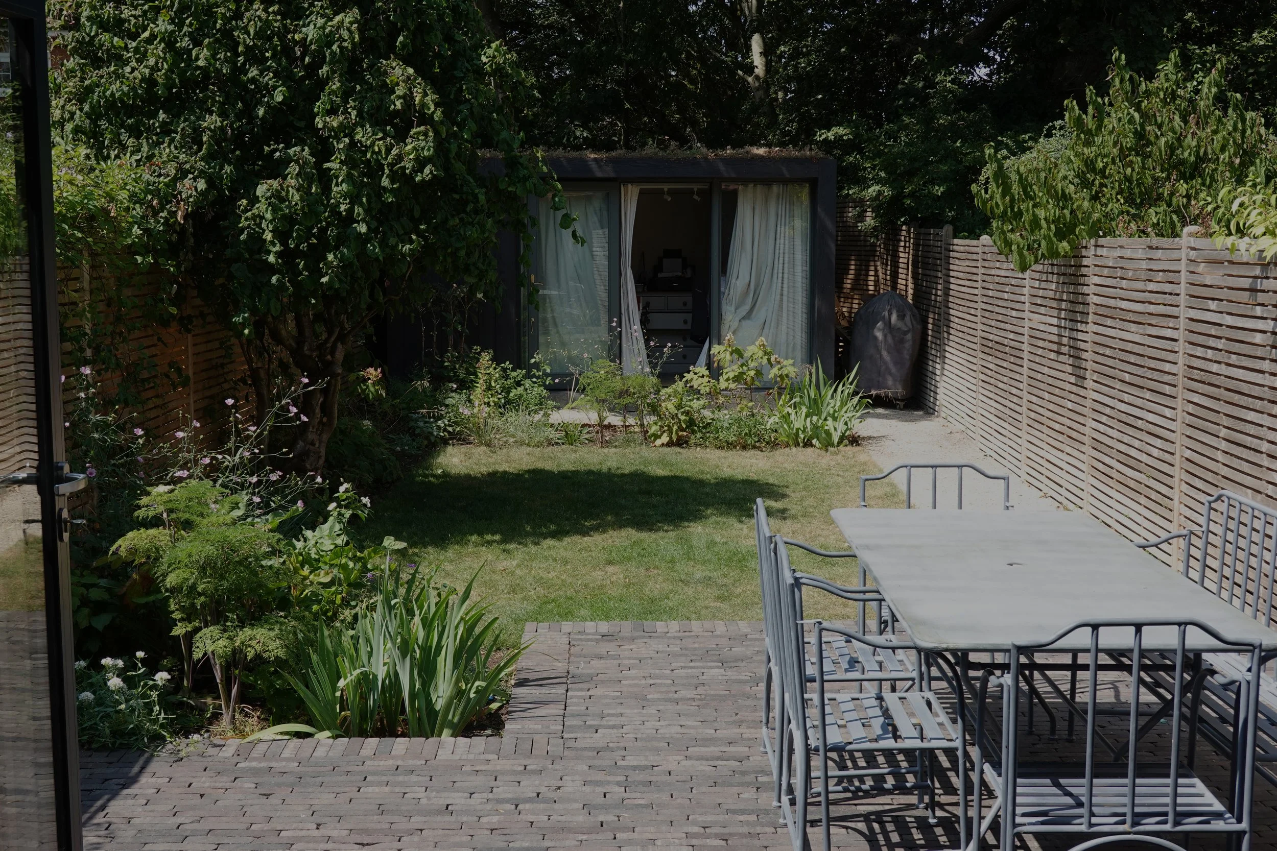 A backyard garden with a brick patio, white outdoor dining table with metal chairs, surrounded by green plants and trees, enclosed by a wooden fence, with a small garden shed in the corner.