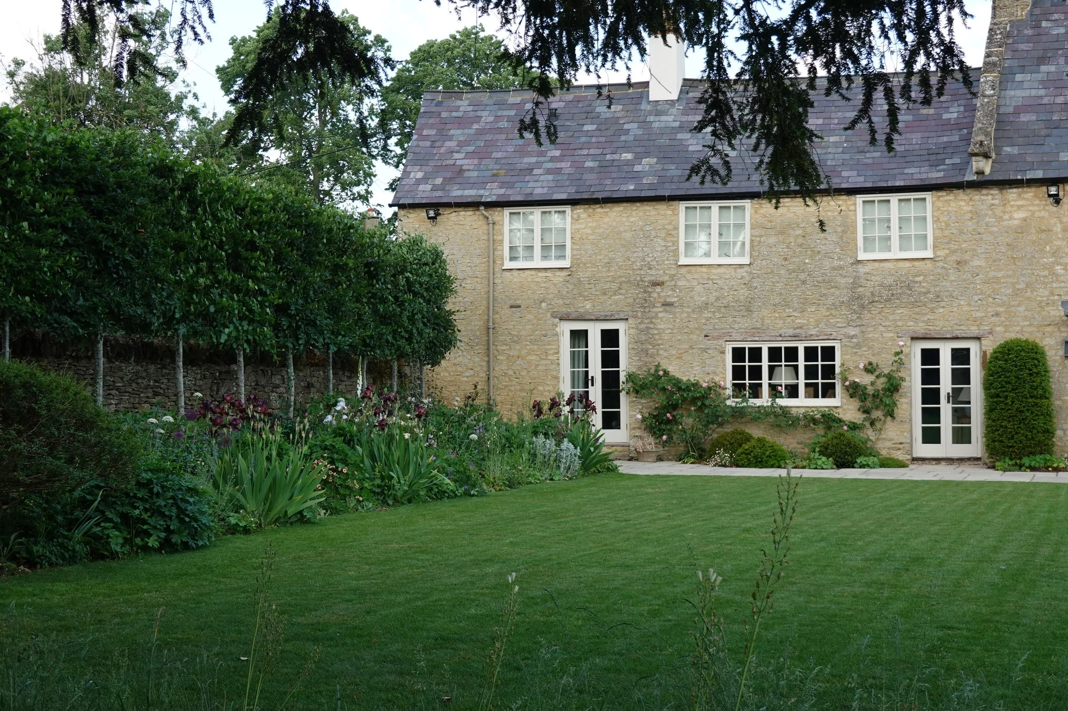 A stone house with a slate roof, surrounded by a lush green garden and trees.