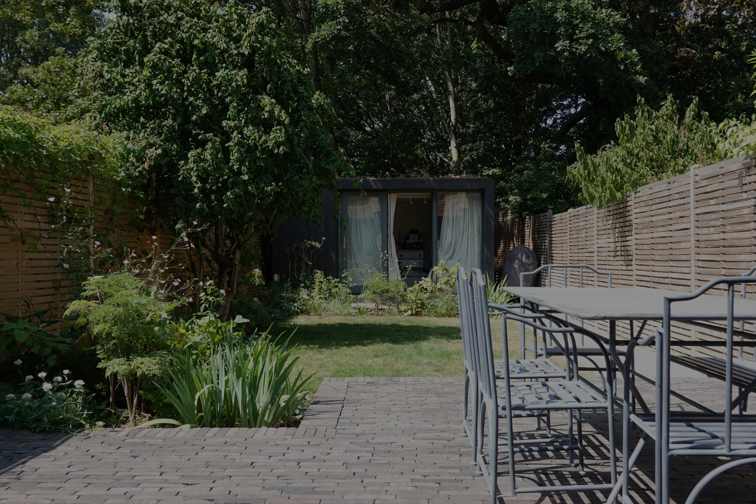 A backyard garden with a wooden fence, green trees, a small garden shed with open doors, outdoor furniture, and a brick patio.