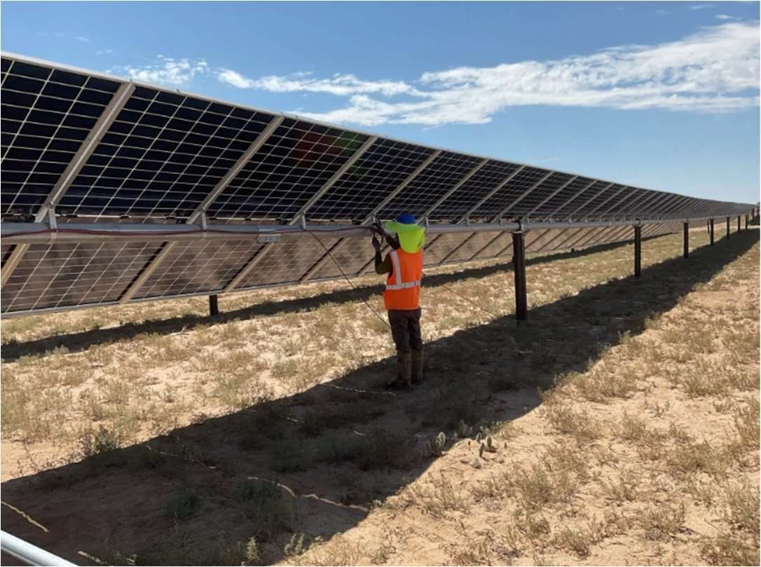 A worker wearing a high visibility orange vest inspects the cabling on the back side of a solar array