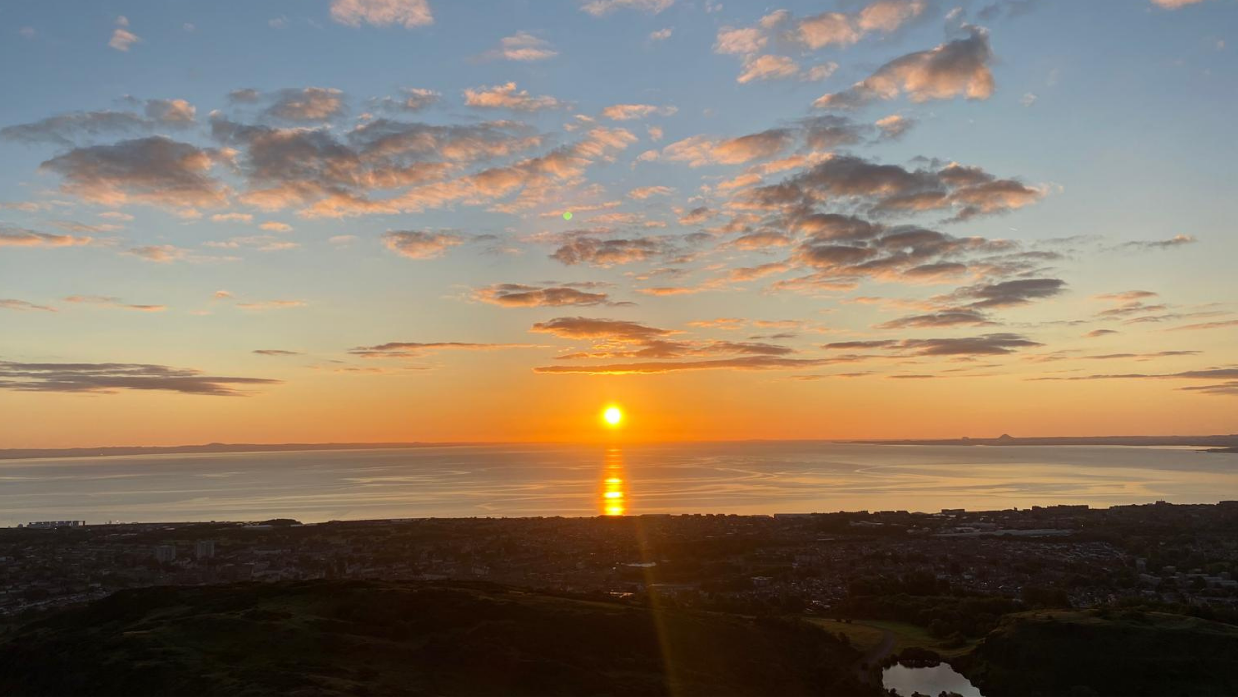 Sunset over a body of water with clouds in the sky and a small hill or land on the horizon.