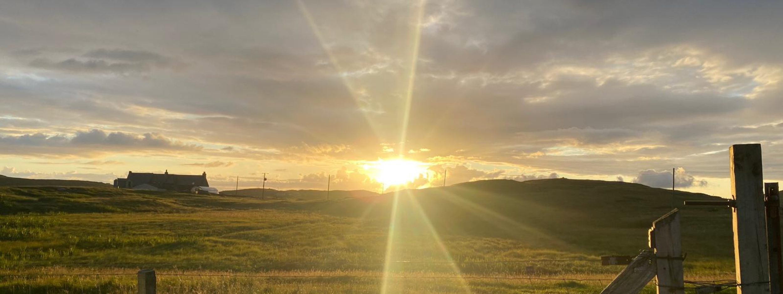 Sunset over a rural landscape with grassy fields, a house in the distance, utility poles, and a partly cloudy sky.