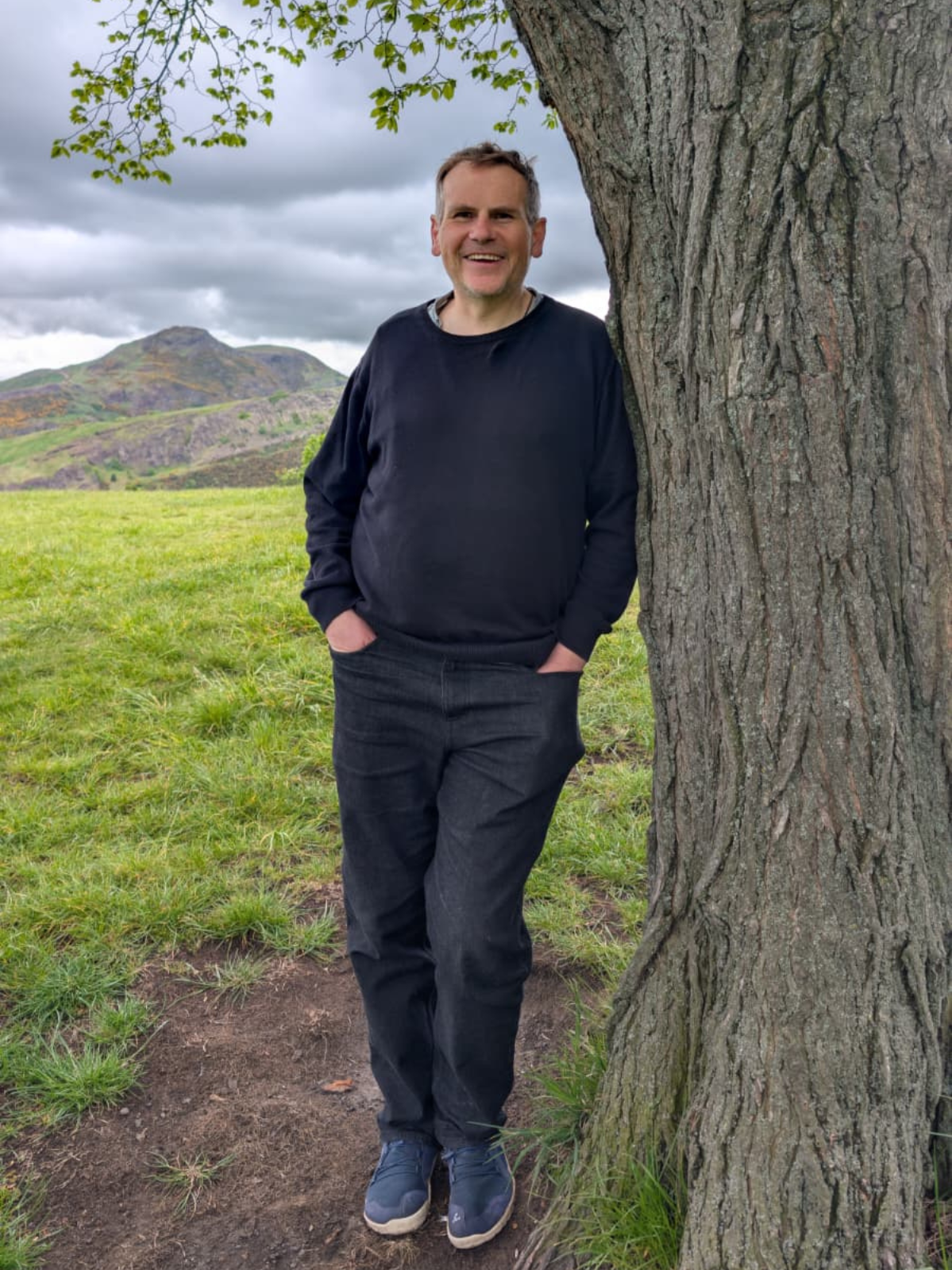 A man standing outdoors beside a large tree with rolling hills and cloudy sky in the background.