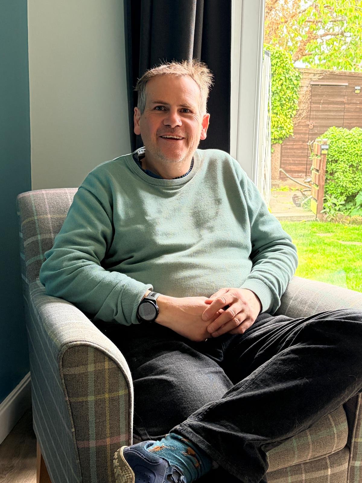 A man sitting in a patterned armchair with a large window behind him showing a garden with green grass and plants.